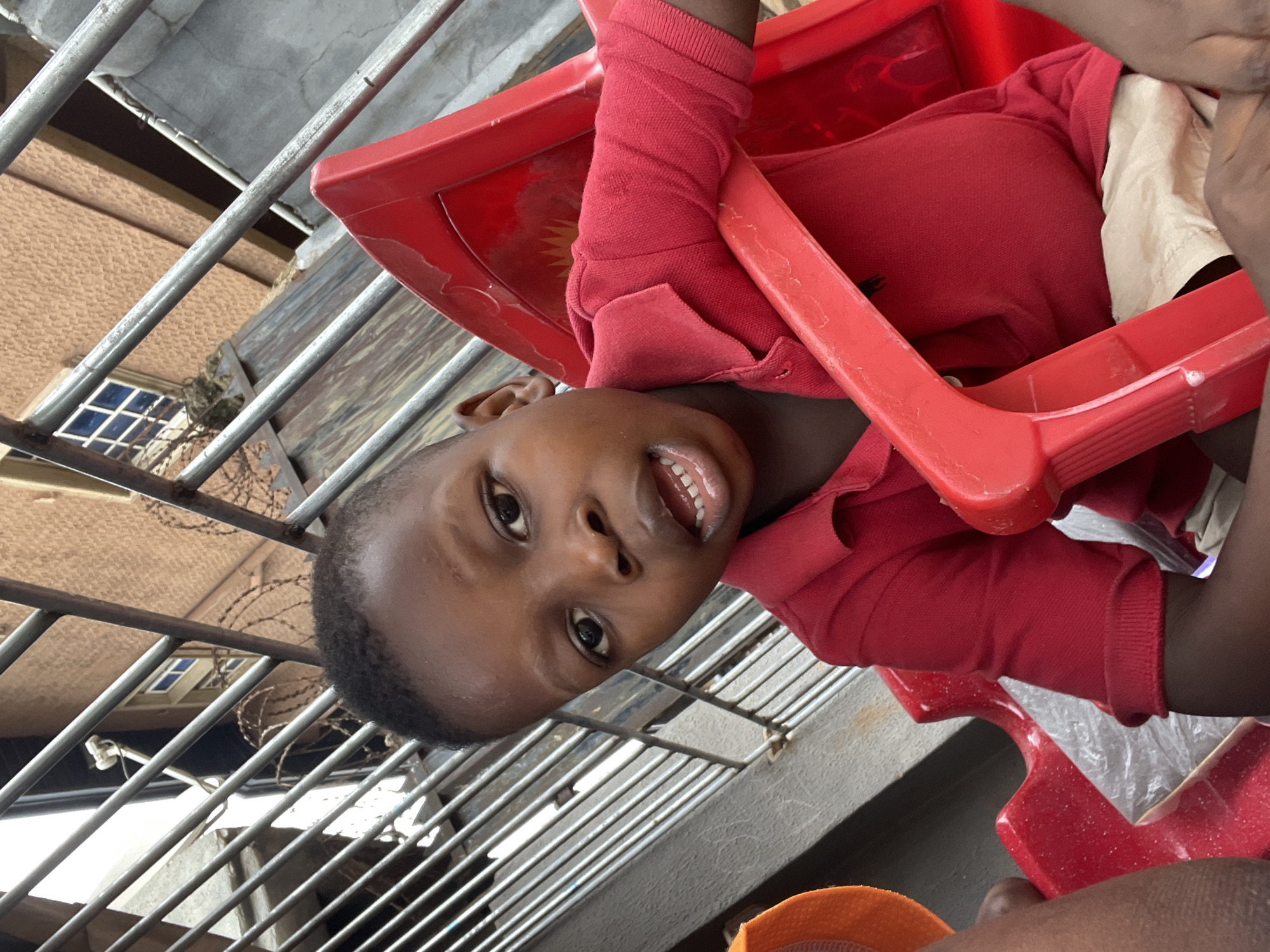 A young boy is seated in a red plastic chair in what appears to be an outdoor or semi-outdoor space in Onitsha, Nigeria. The location is suggested by the background environment and possible building materials. The boy wears a red polo shirt and has a fair complexion. He is looking towards the camera, with his mouth open, perhaps smiling or speaking. In the background, there are vertical metal bars, possibly part of a gate or railing, and a building facade with barred windows and barbed wire can be seen through the bars. The area behind the railing appears to be an urban setting. There is another red plastic chair near the boy and some light-colored fabric is visible in the chair.