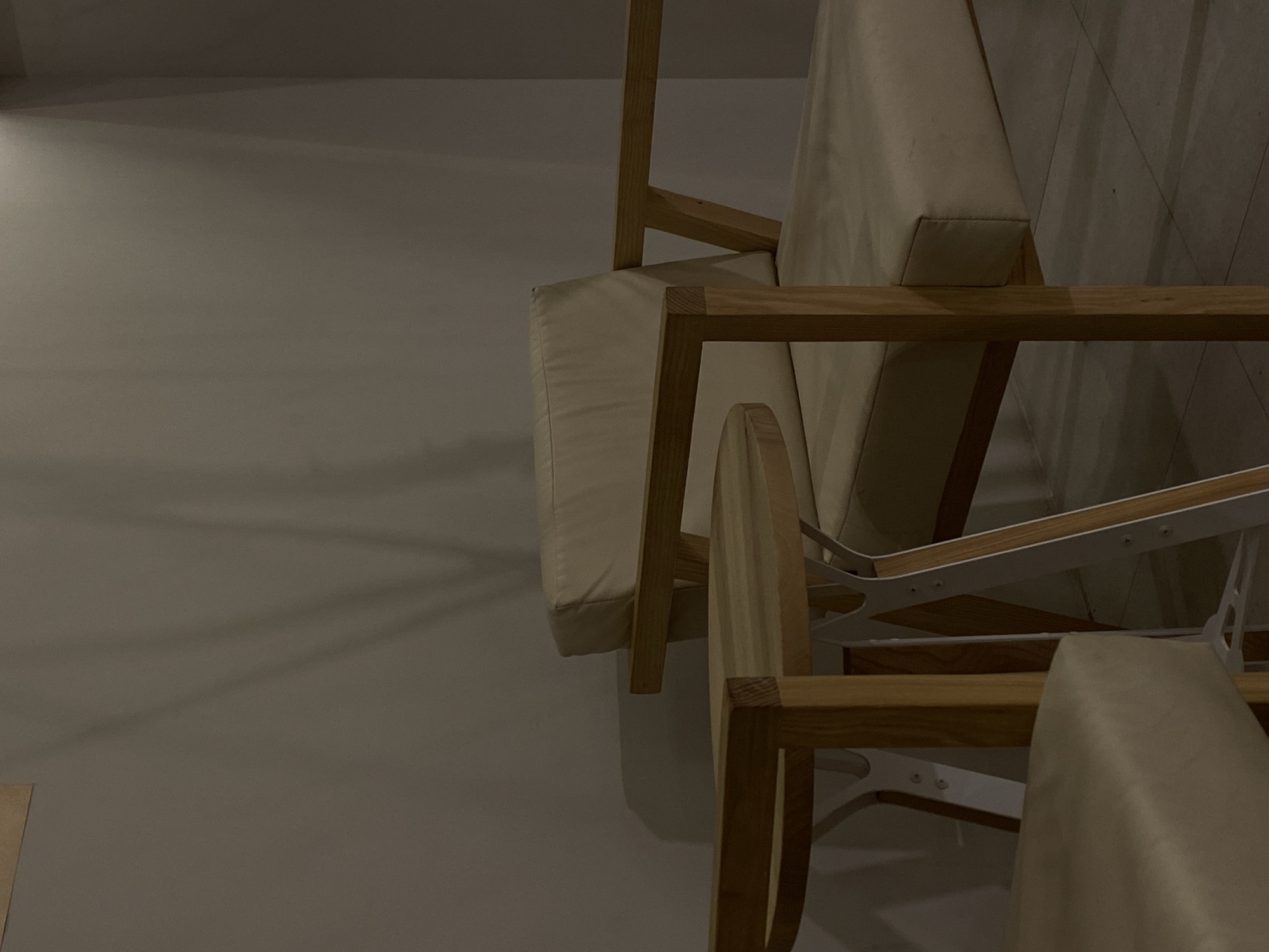 In a room in Vishnyaki, Russia, a light-colored upholstered armchair with wooden armrests and legs sits next to a small wooden side table with a white metal frame. The armchair's cushion and back are a light beige color. The side table has a round wooden top. The floor is tiled in a light color. Shadows from an unseen light source are cast on the wall behind the furniture, creating an abstract pattern.