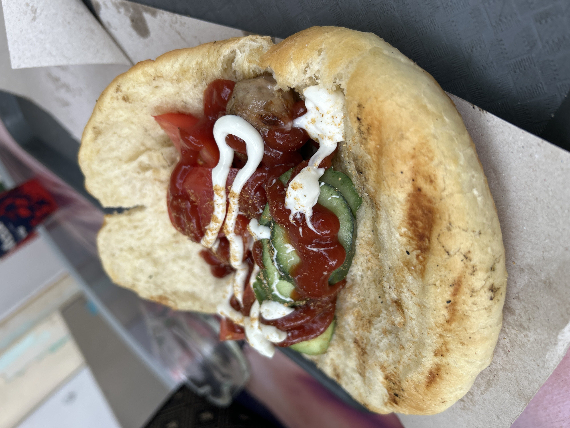 A close-up, eye-level shot shows a grilled bread roll split open and filled with a sausage, sliced cucumbers, chopped tomatoes, ketchup, and mayonnaise. The bread is golden brown with grill marks and a slightly charred appearance. The fillings are arranged somewhat haphazardly, with dollops of white mayonnaise and streaks of red ketchup covering the sausage and vegetables. The entire creation rests on a piece of light brown paper. In the blurred background, there are hints of a dark gray surface and what appears to be a paper cup with a red and blue logo. The overall scene suggests a casual food setting, possibly an outdoor vendor or a street food stall. This image was taken in Obnova, Bulgaria.
