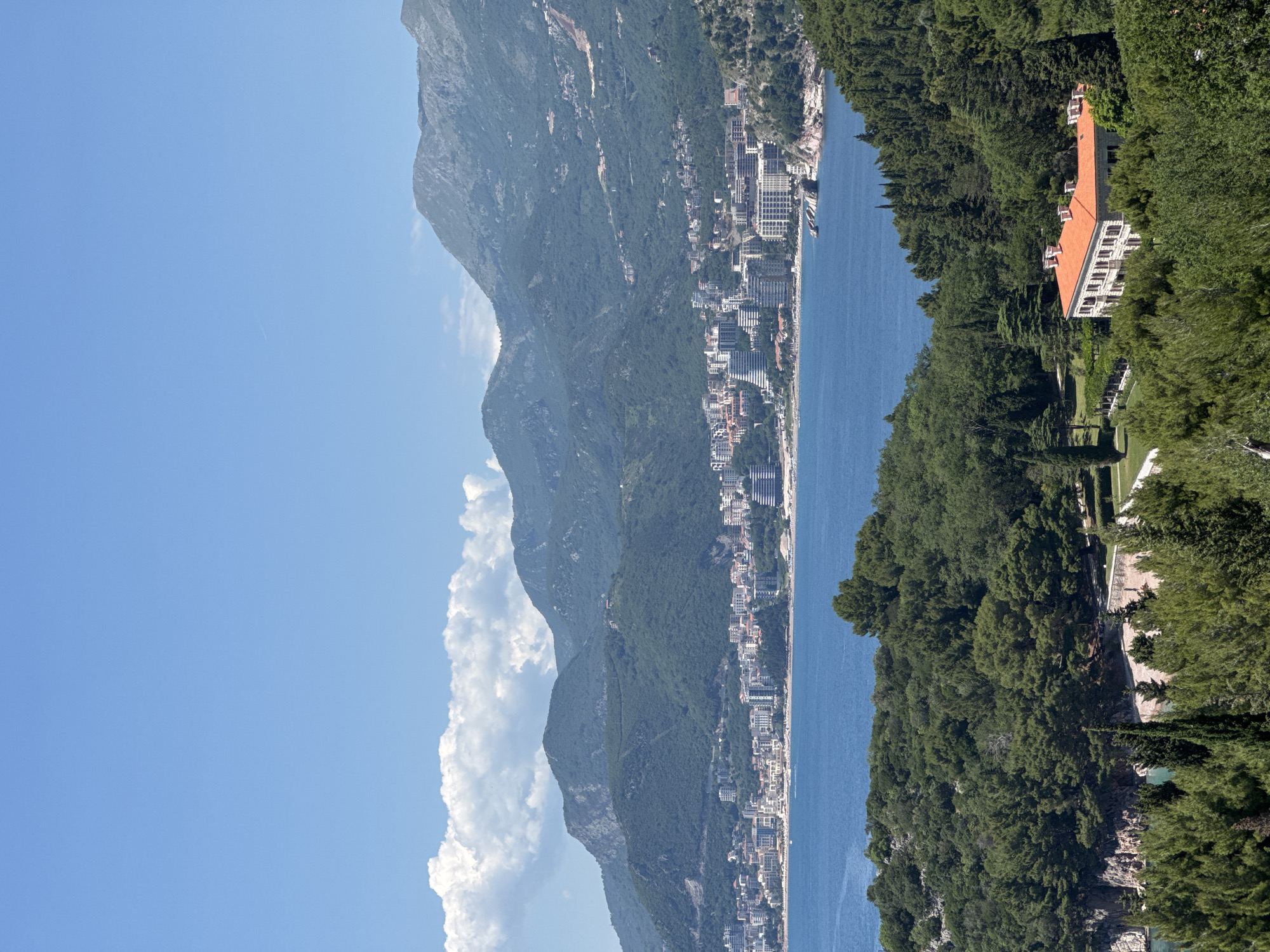 The image shows Sveti Stefan, Montenegro from an elevated perspective. The Adriatic Sea fills the center, bordered by mountains to the left. Buildings line the coast along the mountains. Lush greenery, with a large building with an orange roof, occupies the right side of the frame. The sky is blue with scattered white clouds.
