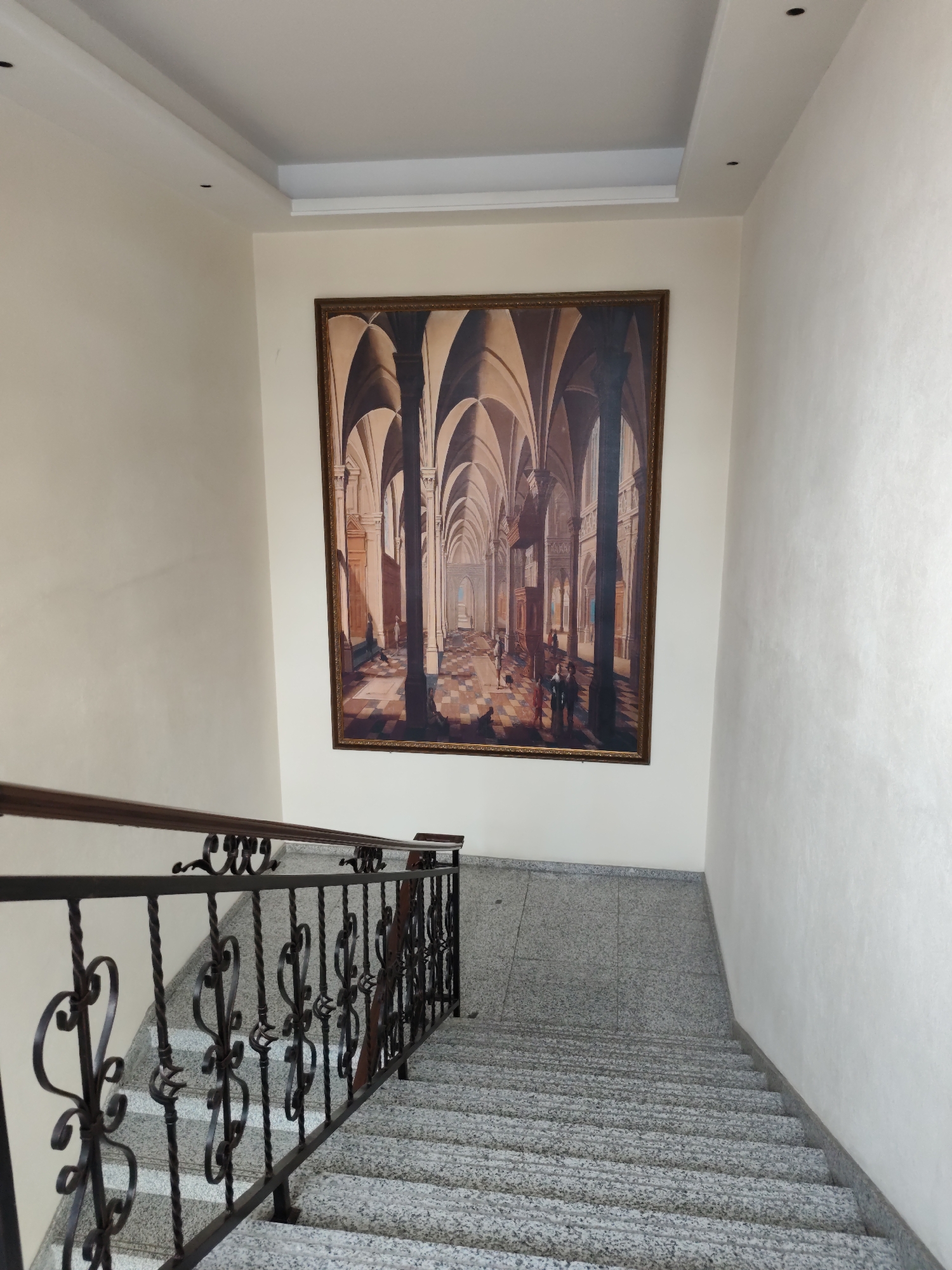 The image shows an interior staircase with a large framed painting on the wall. The painting depicts an architectural scene with arched colonnades and figures walking within. The staircase is made of granite with a dark metal railing featuring decorative scrollwork. The walls are painted a light color, and the ceiling has recessed lighting. The scene appears to be in a well-lit indoor space, possibly a public building or a residential hallway. There is no visible indication that this location is in Aihui, China.