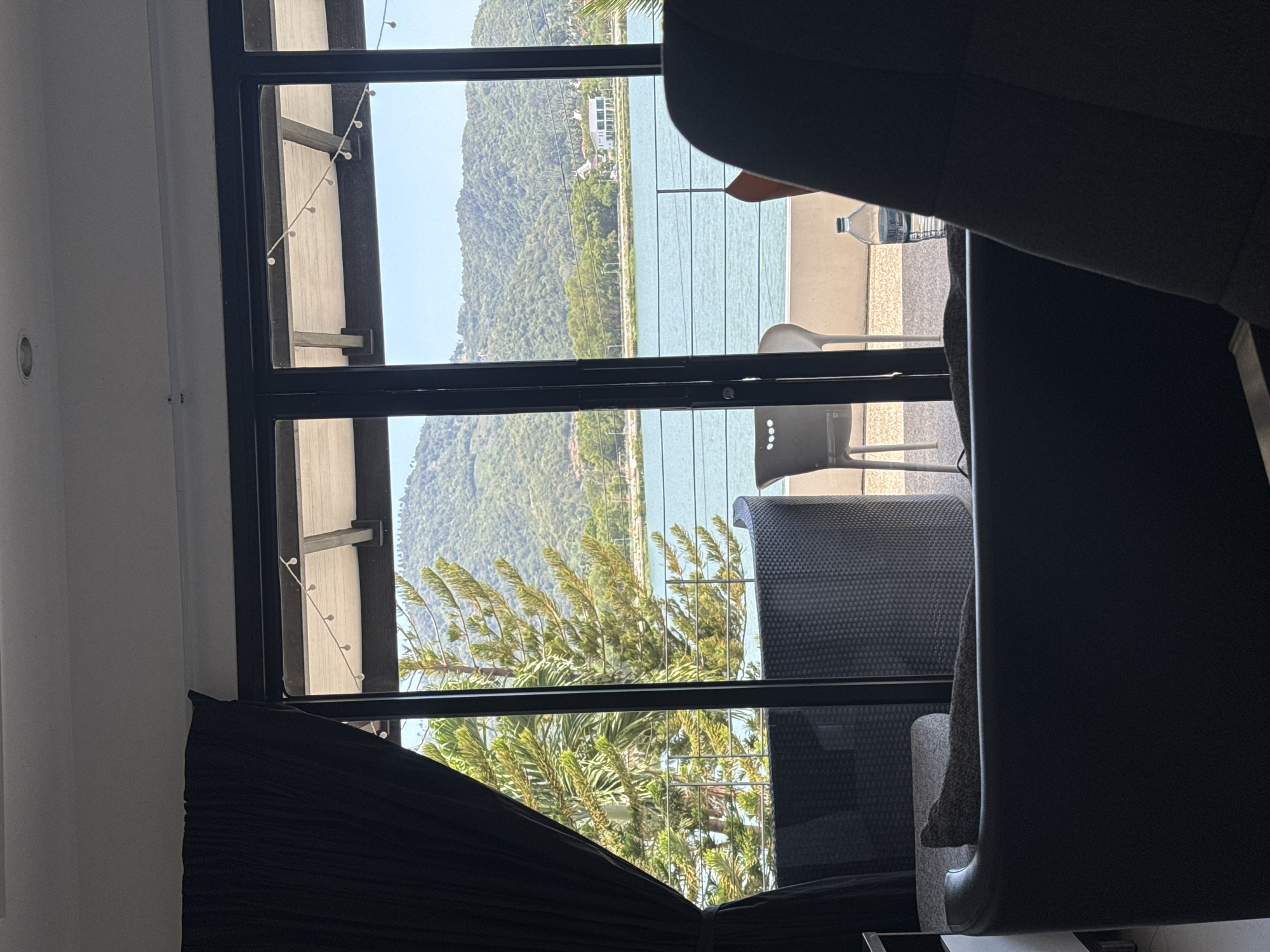 The image depicts a view from indoors looking out through a large glass door and window to a balcony and a body of water with a hilly landscape beyond. The foreground features the back of a dark armchair, partially obscuring a sofa. To the left, a black curtain is drawn. On the balcony, visible through the glass, are pieces of outdoor furniture: a dark wicker armchair and two white plastic chairs. A clear plastic bottle of water with a blue label sits on the balcony floor near the railing. The balcony overlooks a serene body of water, possibly a bay or calm sea, with lush green hills forming the backdrop. A road runs along the edge of the water, with several white buildings and trees visible on the far bank. String lights are attached to the ceiling above the balcony. The overall scene suggests a tranquil setting, likely a resort or residence in a tropical location. The scene is consistent with locations found in Ko Samui District, Thailand, known for its coastal scenery and hilly interiors. There are no people visible in the image.