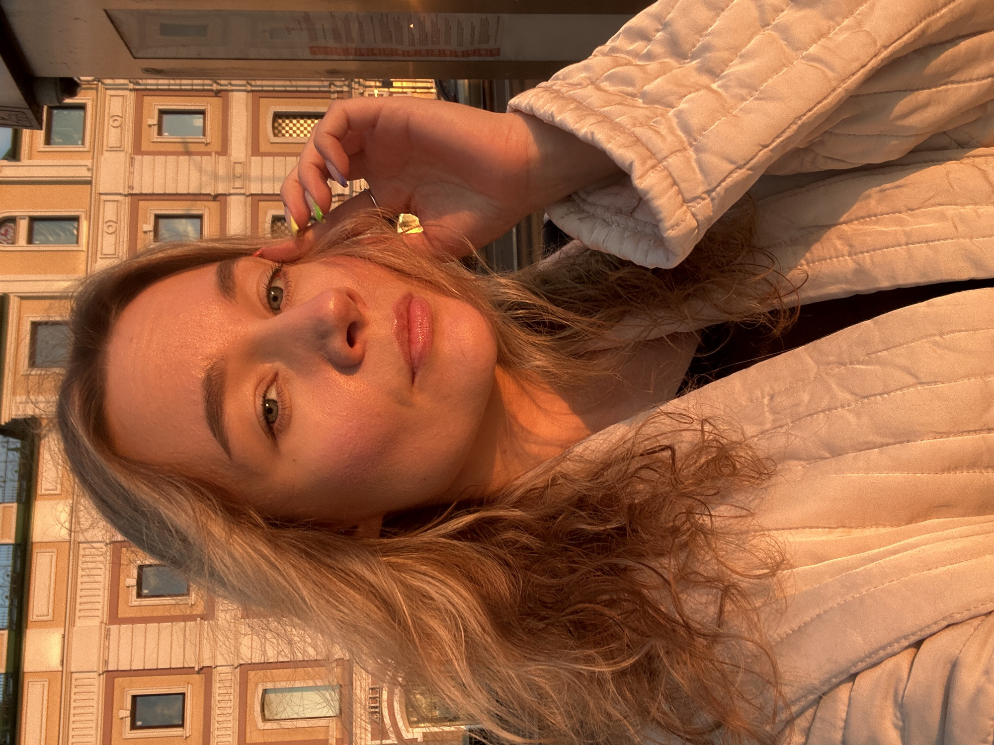 An outdoor selfie shows a young woman with blonde, wavy hair and blue eyes. She is wearing a cream-colored quilted jacket over a dark top. Her right hand is raised to her face, touching her temple. Her fingernails are painted with neon green and purple polish. The woman is illuminated by the warm glow of sunset, casting long shadows and highlighting her features. In the background, a building with several windows and ornate architectural details is visible. The lighting suggests the photo was taken at dusk or dawn. The overall scene is one of casual intimacy and pleasant weather.