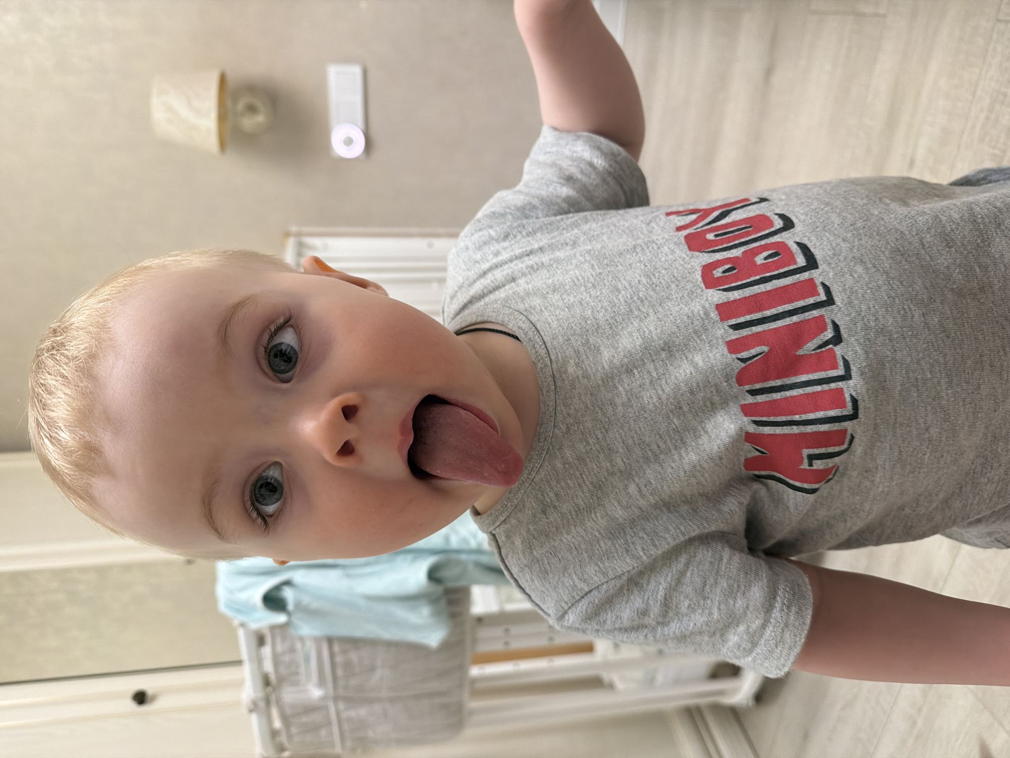 A young child with blond hair and light skin is sticking their tongue out, looking directly at the camera. The child is wearing a gray t-shirt with the word "MINIBOY" printed in red and black. The background includes a wall with a light fixture and a control panel. There is a dresser or cabinet visible, and a light blue piece of clothing is draped over it. The floor appears to be light-colored wood or laminate. There is no information in the image to confirm it was taken in Votkinsk, Russia.
