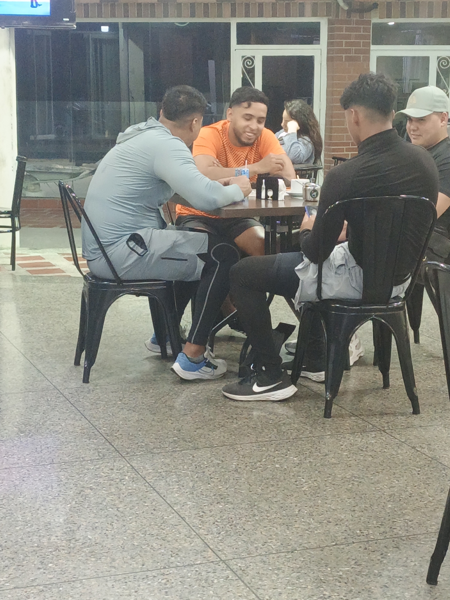 Four young men are seated around a small, dark-colored table inside a restaurant or cafe. The location is suspected to be in Tinaquillo, Venezuela. The man on the left is wearing a light blue hooded jacket and gray shorts. The man across from him is wearing an orange shirt and smiling. The man to the right of the smiling man is wearing a black shirt. The fourth man wears a baseball cap. They are seated in metal chairs with open lattice backs. On the table are assorted items including cups, napkins, and possibly a phone. The floor is a speckled gray tile. Large windows are in the background.