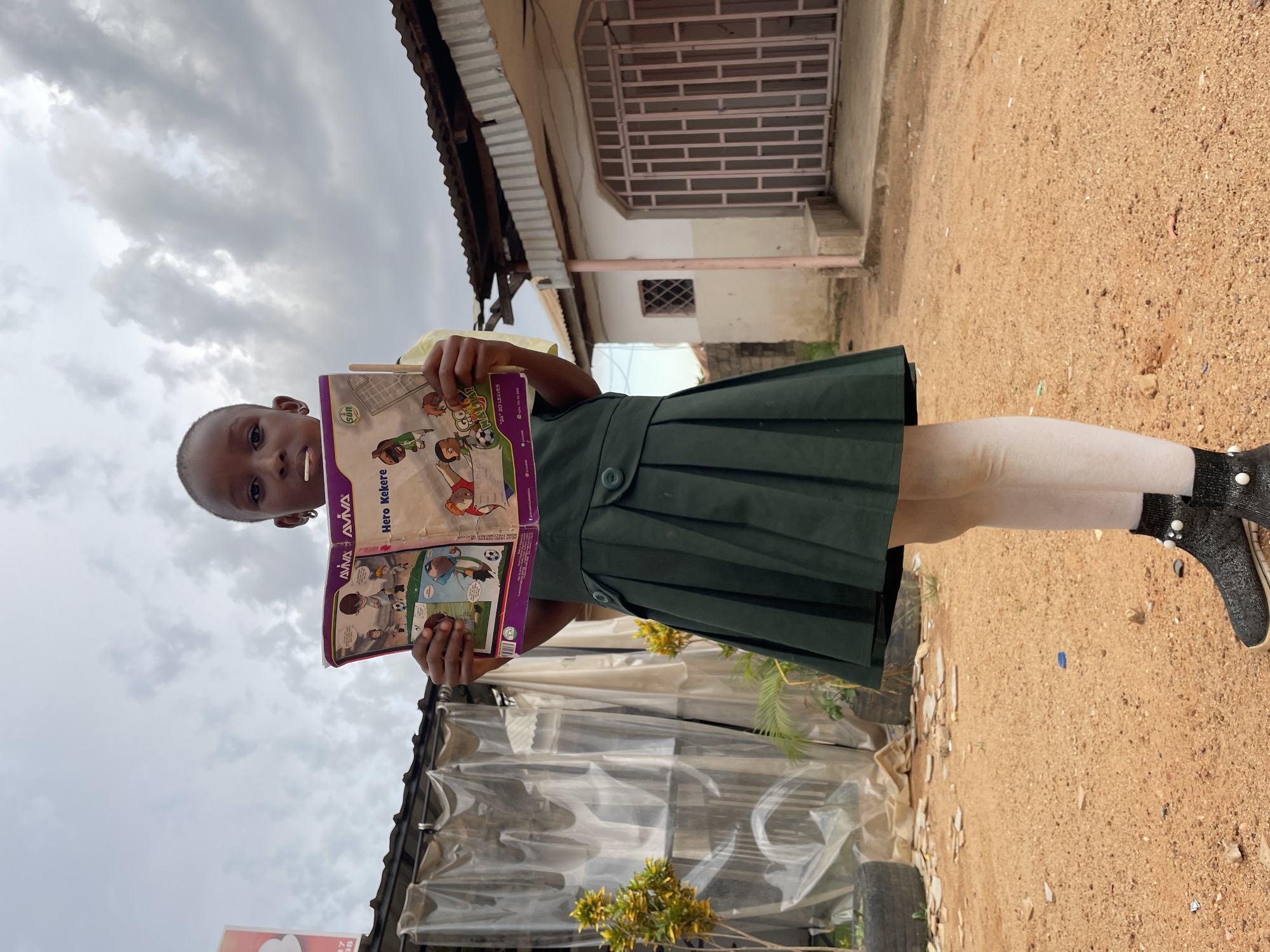 A young girl stands in front of a residential building, holding a comic book open towards the camera. The comic book appears to be titled "Hero Kekere." She wears a green pleated school uniform dress with tights and dark shoes. The ground is dirt. The background shows a building with barred windows and a corrugated metal roof overhang. The location is likely in Jos, Nigeria.