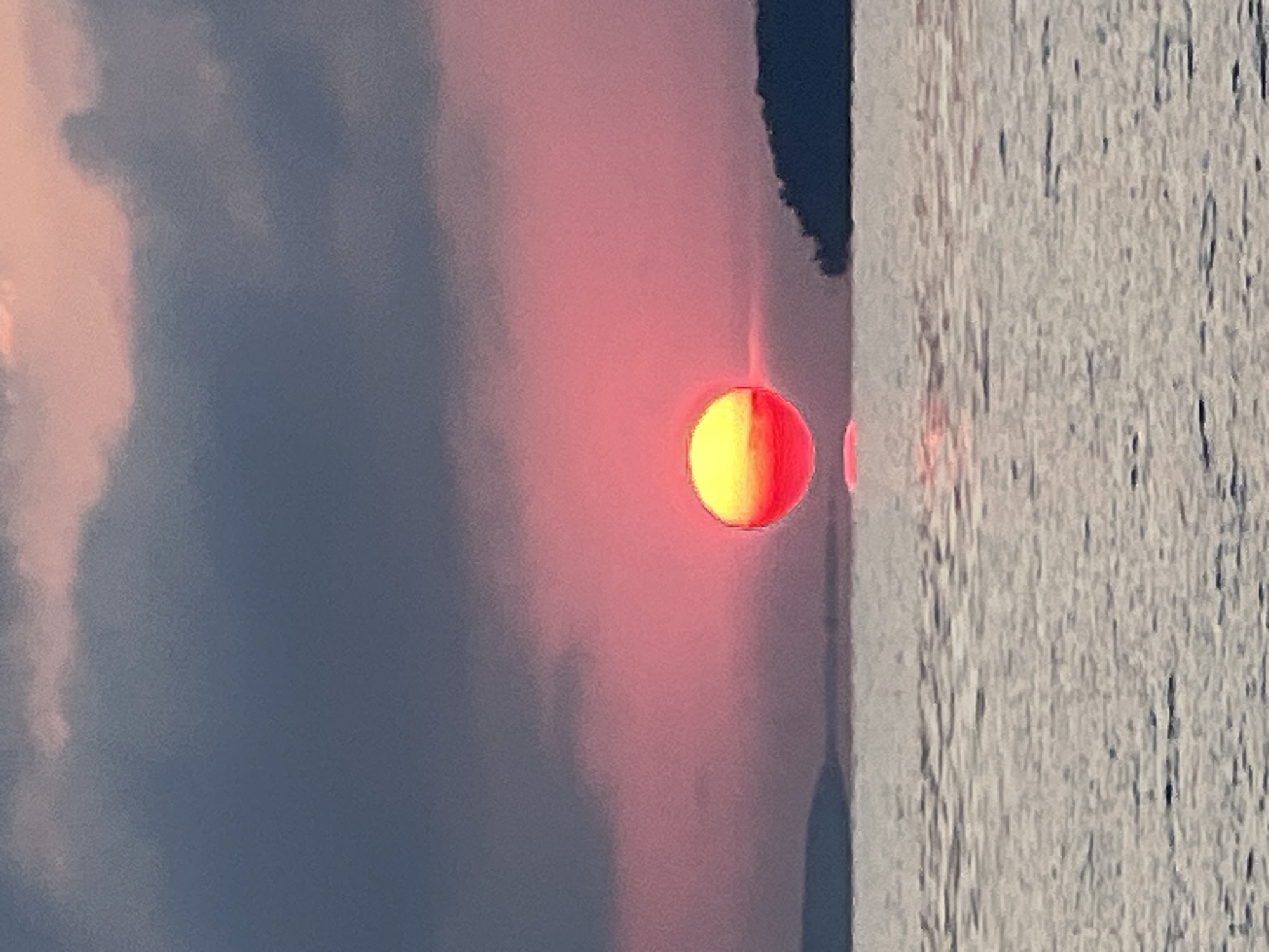 A sunset is depicted over a body of water, likely a lake or sea, near Novopichugovo, Russia. The sun, a vibrant orb of yellow and red, is positioned low on the horizon, casting a warm glow across the sky, which is painted with shades of pink and dark gray clouds. The water's surface reflects the colorful sky, creating a shimmering effect. On the right side of the image, a dark silhouette of land, possibly an island or shoreline, is visible against the horizon. On the left, a faint outline of land can also be seen. There are no people or animals visible in the scene. The overall impression is one of natural beauty and tranquility.