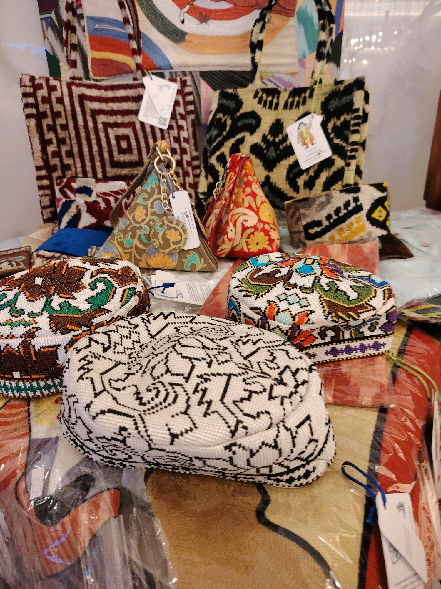 The image depicts a collection of decorative textiles, likely handcrafted items, displayed on a surface covered with a patterned cloth. In the foreground, several beaded caps are prominently featured. One cap on the left is predominantly brown with intricate green and white beadwork forming geometric patterns. To its right, a cap with a white base is adorned with black geometric beadwork. In the center, slightly behind these caps, is another cap with white as its base, intricately decorated with colorful beadwork, including floral motifs in shades of green, brown, blue, orange, and purple.
Behind the caps, several bags and pouches are visible. Two larger tote bags are placed towards the back. The one on the left has a maroon and white geometric pattern, while the one on the right features a black and tan animal print. In front of these, several smaller, triangular pouches or bags are arranged. One is a muted olive green with embroidered floral patterns in gold, blue, and teal. Another is a vibrant red with yellow floral embroidery. There are also smaller rectangular pouches with woven patterns.
The overall impression is one of a retail display, possibly in a market or shop. The items are colorful and intricately designed, suggesting a focus on traditional crafts. The presence of tags on some of the items indicates they are for sale. The context of Konigil, Uzbekistan, suggests these items might be traditional Uzbek handicrafts, such as suzani embroidery or beadwork, which are popular souvenirs and decorative items from the region. The lighting in the scene is somewhat diffused, and the background is softly blurred, focusing attention on the displayed goods.