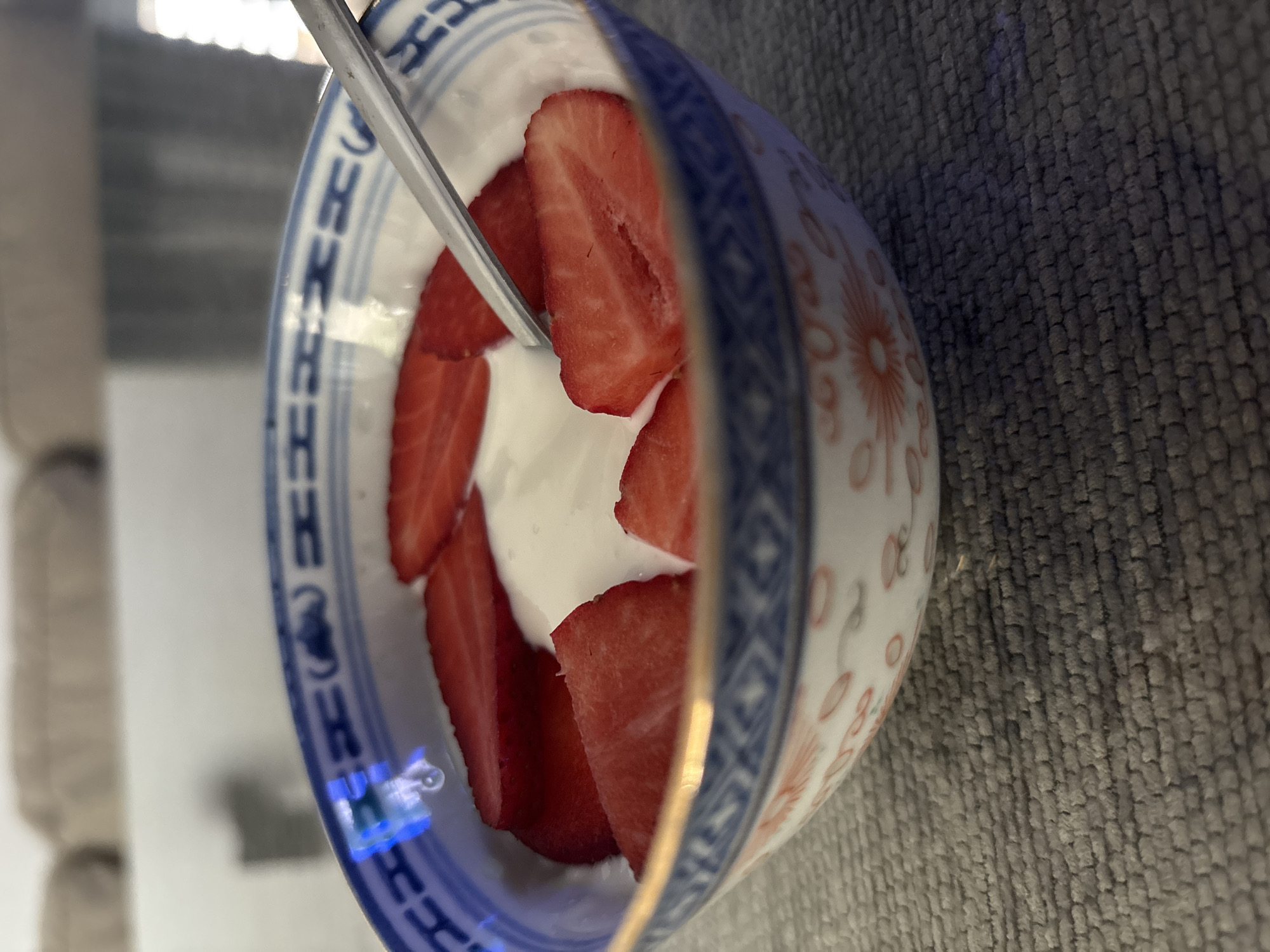A close-up, slightly rotated image depicts a porcelain bowl containing yogurt and sliced strawberries. A spoon is partially visible within the bowl. The bowl features a traditional blue and white design along its rim, with a red floral pattern on the side. The background shows a gray, textured surface and a partially visible interior space. The location is not discernible from the image. There is no information related to the Urbanización Encinares De Alcolea.