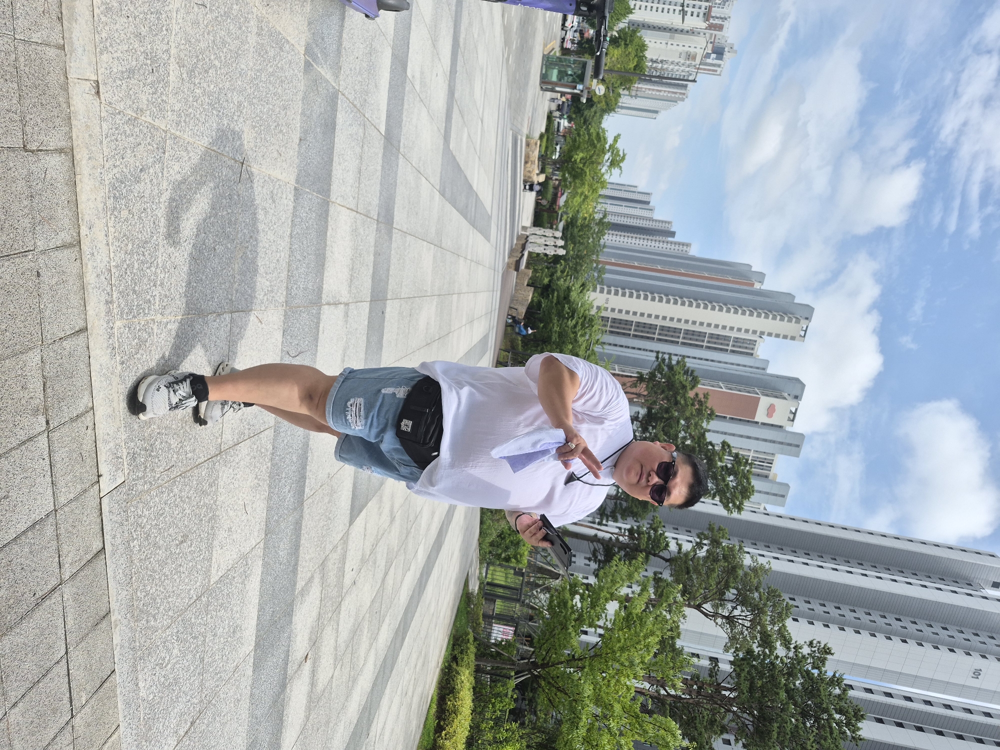 A medium shot captures a man standing on a paved walkway in Ansan, South Korea, with a backdrop of tall apartment buildings. The man, wearing sunglasses, a white t-shirt, and ripped denim shorts, holds a small white cloth in his left hand and a black tablet in his right. He is wearing a black fanny pack around his waist and grey athletic shoes. The walkway is made of large, light-colored square tiles. In the background, several modern, high-rise apartment buildings dominate the skyline, interspersed with green trees. The sky above is blue with scattered white clouds. To the left of the frame, a purple electric scooter is partially visible. Several other people can be seen in the distance, some walking and others sitting. The overall scene depicts a bright, sunny day in a residential urban area.