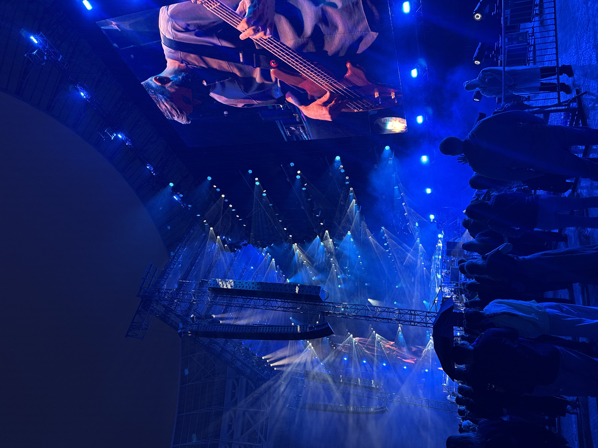 The image depicts a live music concert at night, likely an outdoor event given the open-air stadium seating visible in the background, illuminated by intense blue stage lighting and pyrotechnics. A large screen displays a close-up of a male musician playing a bass guitar, his focus directed downwards. In the foreground, a crowd of attendees are gathered, silhouetted against the bright stage lights. One individual in the crowd holds a black umbrella, suggesting possible rain or a desire for shade from the lighting. The overall atmosphere is energetic and immersive, with fog or smoke machines contributing to the dramatic visual effects. The scene is characterized by a strong blue color palette due to the lighting. The presence of a large stadium suggests a capacity for thousands of spectators. The context implies a well-established venue capable of hosting major musical acts.