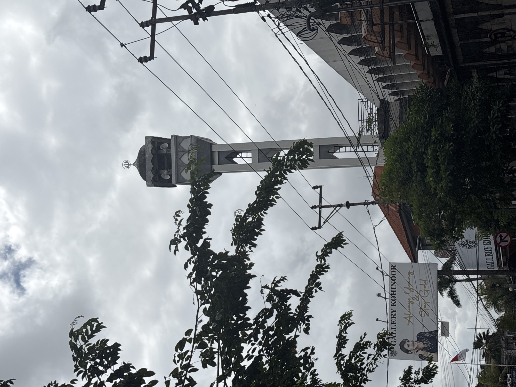 The image captures a cloudy sky over a city scene in Denpasar, Indonesia. In the mid-ground, a tall, slender minaret of a mosque rises, topped with a dome and the Arabic word for Allah. The minaret has multiple levels with decorative grey and white accents and open archways. In the foreground, a large billboard advertising "Gallery Kohinoor" is visible, featuring a woman and the slogan "Always Young & Stay Gold." Adjacent to the billboard, a red and white Indonesian flag flies from a flagpole. Electrical wires crisscross the sky, attached to utility poles. To the right of the minaret, a building with a distinct arched facade and stacked terracotta roof tiles is visible. Trees with dense green foliage are present, some obscuring parts of the buildings and the billboard. In the lower right, a circular red and white sign with a black arrow indicates a traffic direction. The overall impression is a street view with religious and commercial elements under an overcast sky.