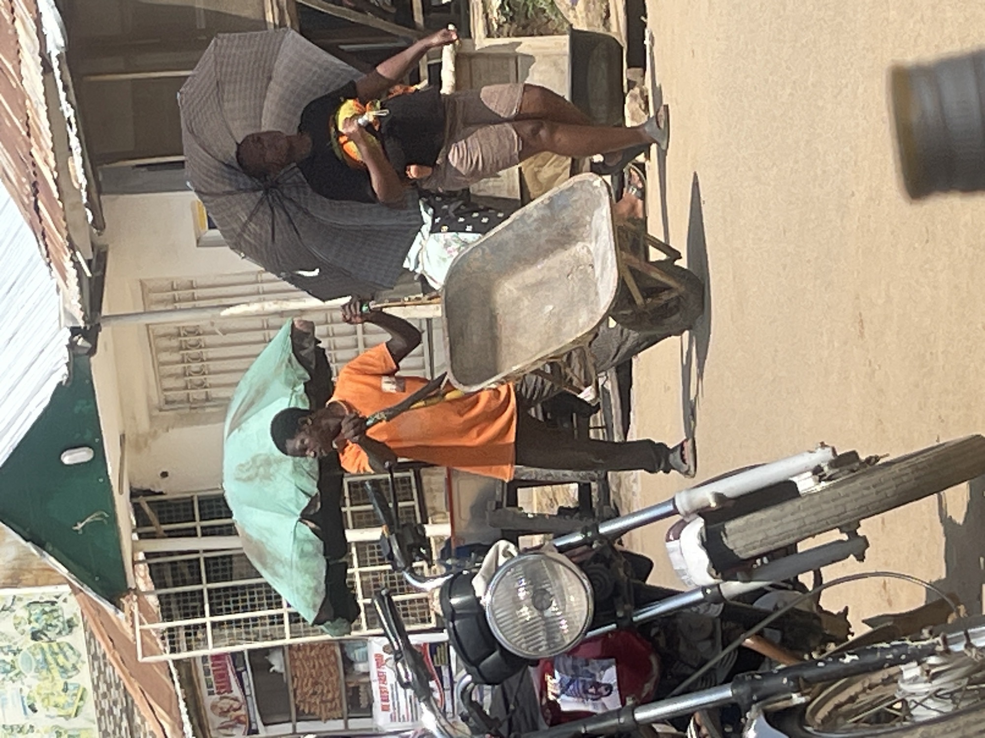 In Abuja, Nigeria, a person in an orange shirt is pushing a wheelbarrow. Another person with a black shirt and shorts is holding an umbrella and a baby. A motorcycle is parked in the foreground. The background shows buildings with corrugated metal roofs, suggesting a market or street scene. The ground is dusty.