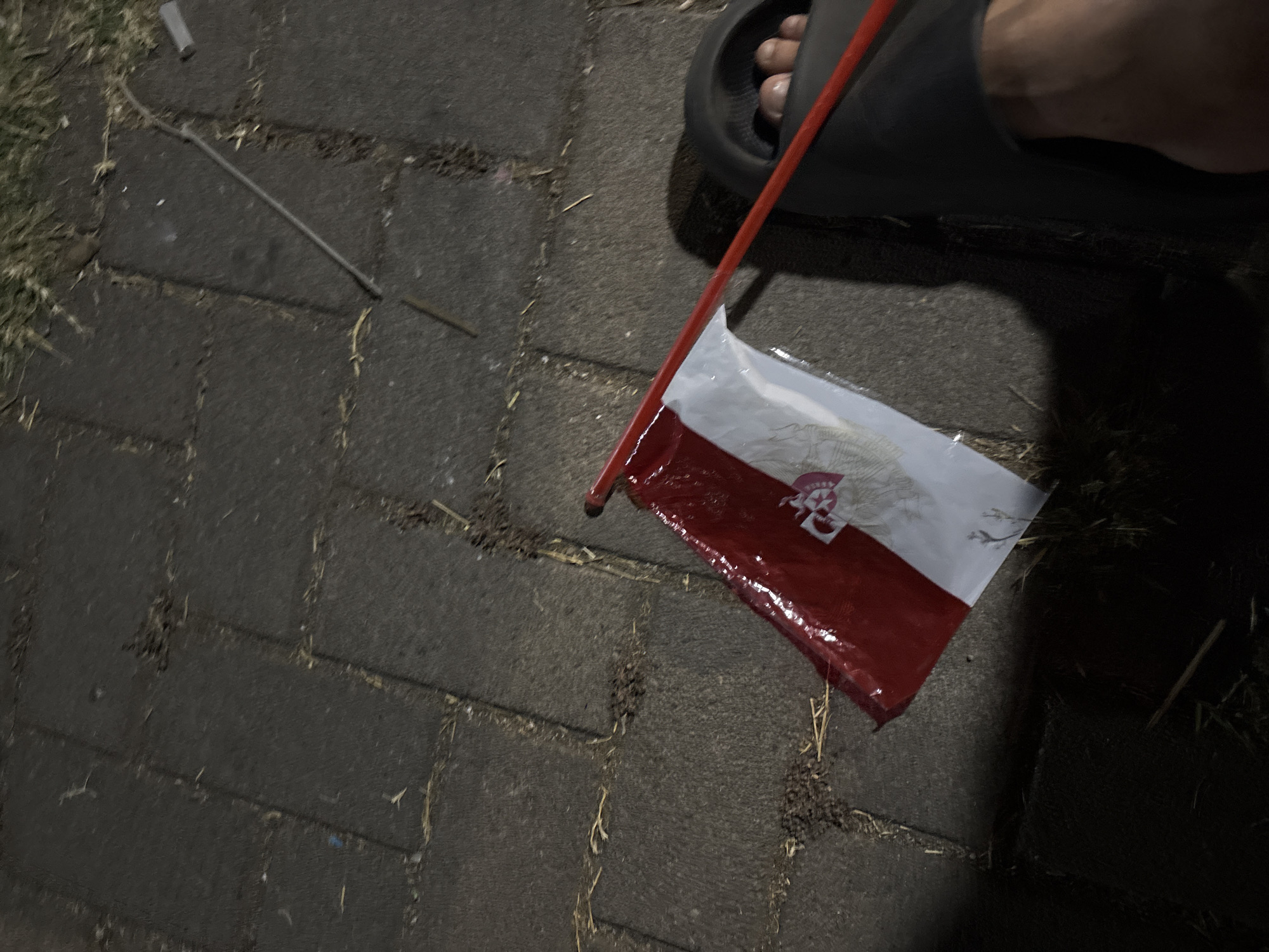 A close-up shot shows a person's bare foot wearing a black slide sandal, positioned on a grey paved ground made of square tiles. The sandal is slightly lifted, revealing the foot. A small flag with red and white stripes and a crest in the center is held by a thin red pole, diagonally positioned across the frame. The flag appears to be wet and slightly crumpled. The ground is littered with small debris, possibly dried grass or leaves. In the background, a thin metal rod is visible lying on the ground, extending upwards and to the right. The scene is dimly lit, suggesting it is nighttime or a shaded outdoor area. The overall impression is a casual, possibly celebratory, moment captured in an urban or semi-urban outdoor setting.