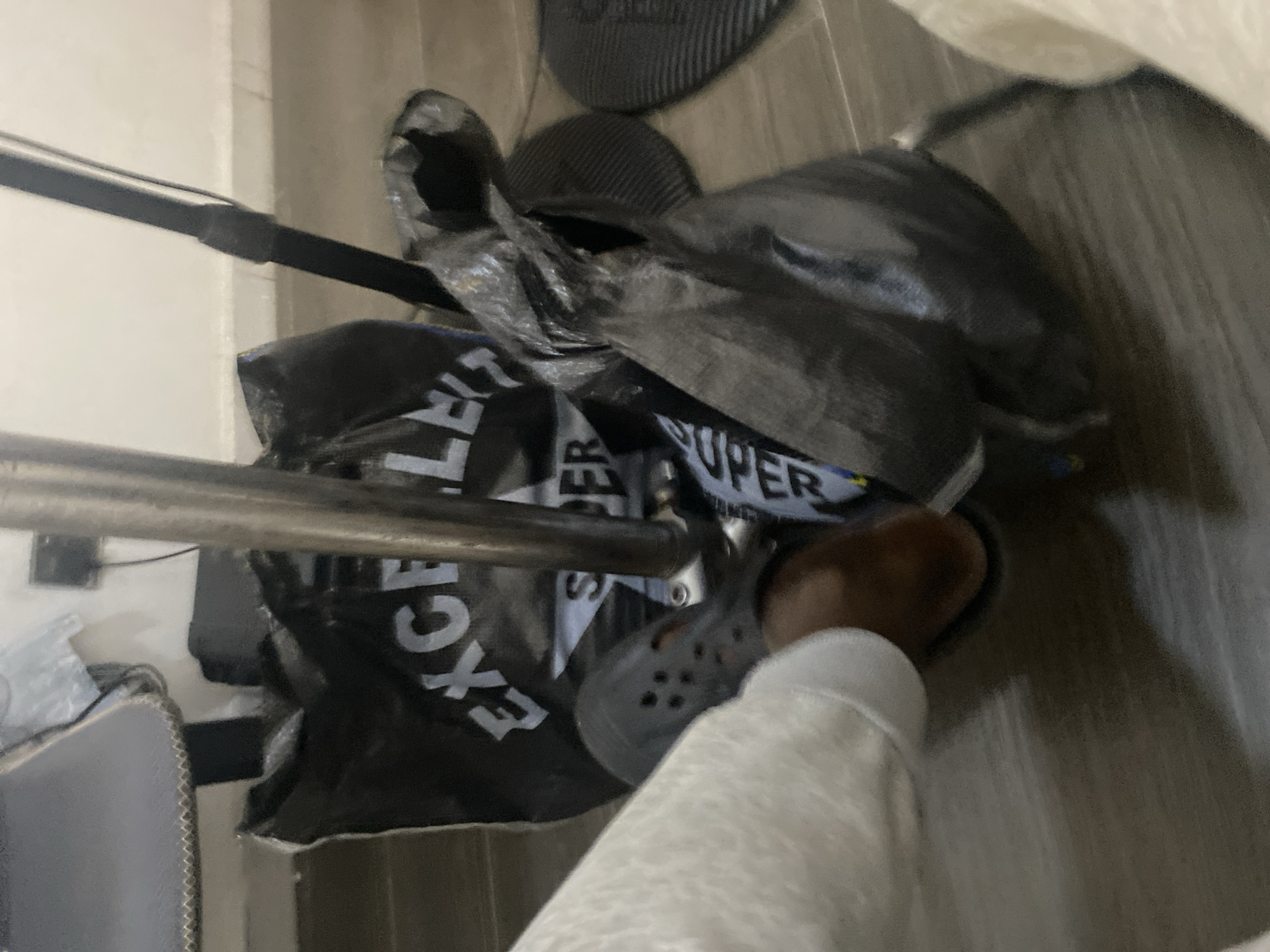 The image, captured from a top-down perspective, depicts an indoor scene with a person's foot and several items on the floor. In the lower right and center, a person's right foot is visible, wearing a light grey or white sock and a dark grey Croc-style shoe.
The central and right portions of the frame are occupied by a pile of dark-colored plastic bags, which appear to be full or crumpled. White text is visible on some of these bags; notably, the word "EXCEL" is clearly readable on one bag, and "SUPER" is seen on another.
In the upper background, resting on a light-colored wooden or laminate floor, are two dark, round objects with ribbed textures, possibly floor mats. A metallic, cylindrical pole extends horizontally across the mid-left of the image, partially obscuring some of the bags. Above this pole, a darker, thinner bar is also visible. The background on the far left shows a light-colored wall with some minor dark marks or wires. A small part of a light grey object with a stitched edge, possibly a mousepad, is just visible on the bottom left. The setting appears to be an everyday indoor environment.