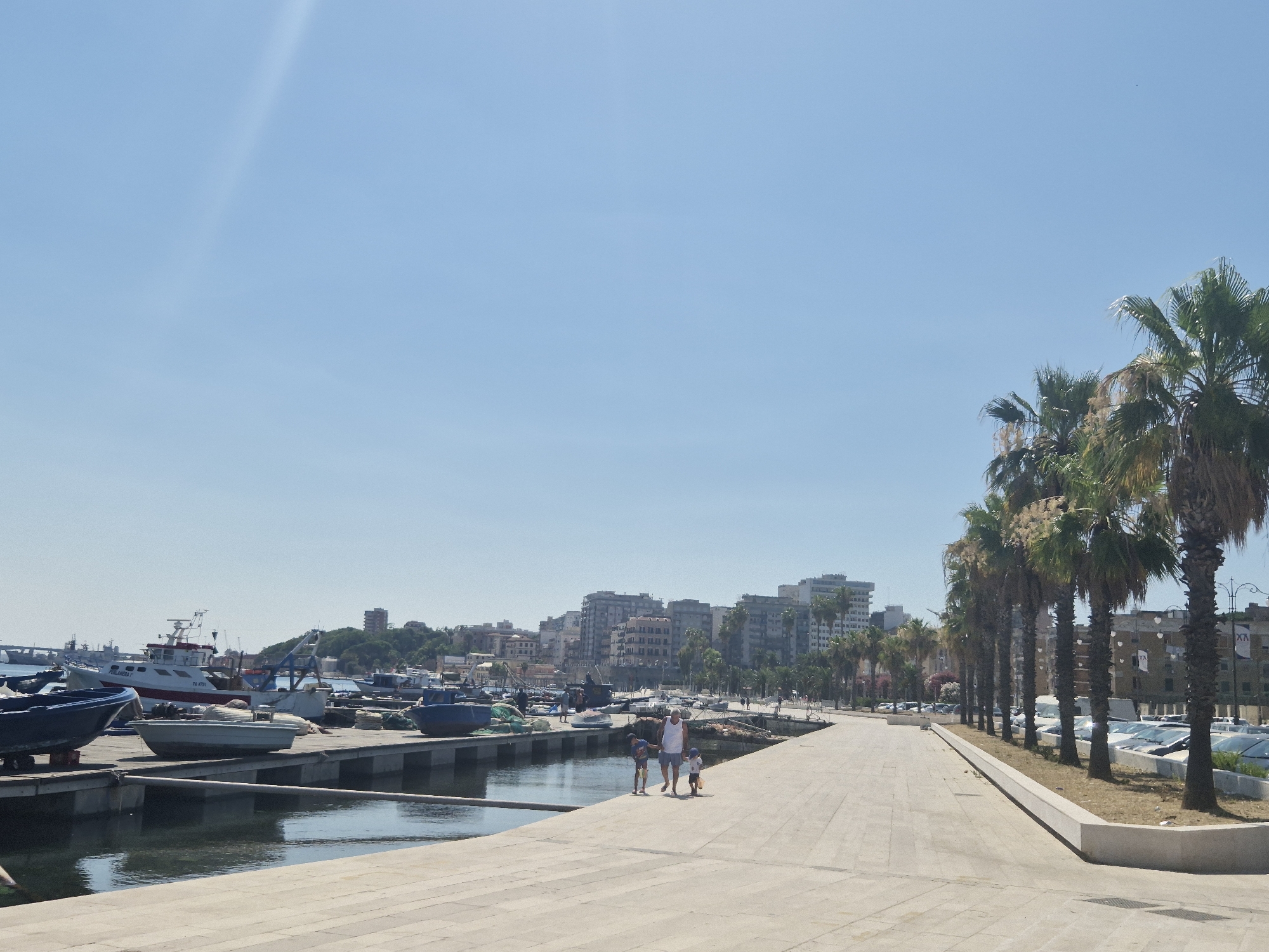 The image shows a waterfront promenade in Taranto, Italy, on a bright, sunny day. The promenade is paved with light-colored tiles and runs along a harbor filled with various boats, mostly fishing vessels. In the foreground, two children and an adult, possibly a father and his children, are walking along the promenade, with the children appearing to be playing. To the right of the promenade, a row of palm trees stands between the walkway and a parking lot filled with cars. In the background, a city skyline with numerous buildings, including some tall structures, rises behind the harbor and palm trees. The sky is clear and blue, with a faint streak of light in the upper left quadrant. The overall scene depicts a pleasant, leisurely day at the harbor in a coastal Italian city.