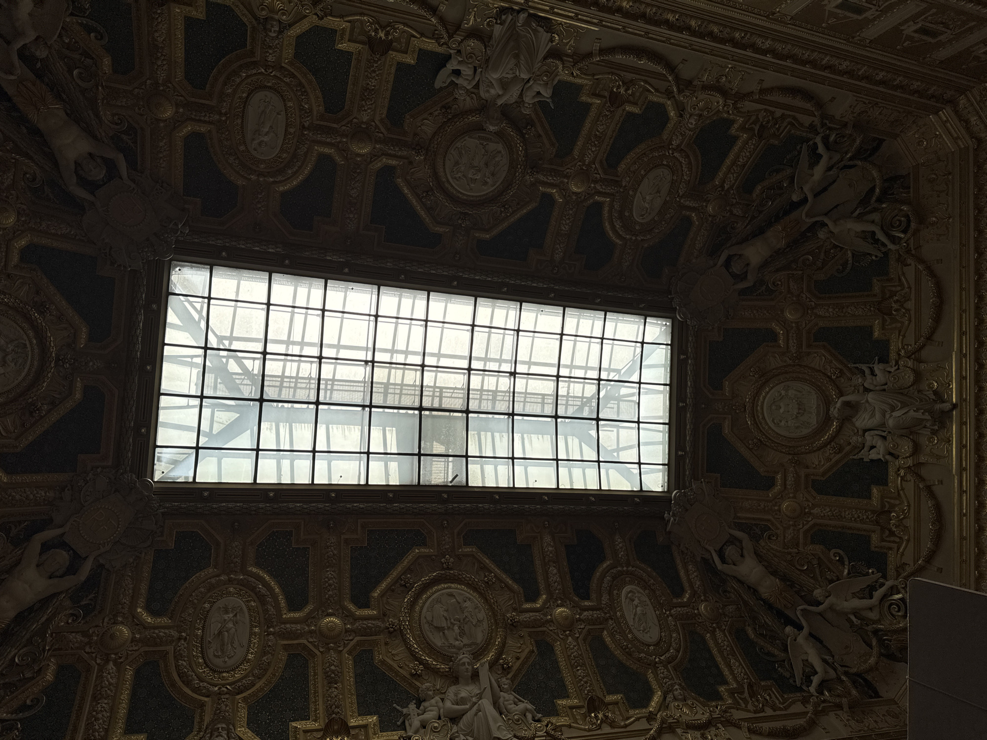 This is a low-angle, full shot of an ornate, gilded ceiling with a large, rectangular skylight in the center. The ceiling is heavily decorated with intricate gold-colored reliefs and medallions against a dark background. The reliefs depict classical figures and mythological scenes. The skylight is framed by a grid of metal bars and is letting in diffused natural light. The overall impression is one of grandeur and historical significance. This ceiling is likely located in a prominent Parisian museum or palace, such as the Louvre Museum or the Palace of Versailles, given its opulent design and architectural style.