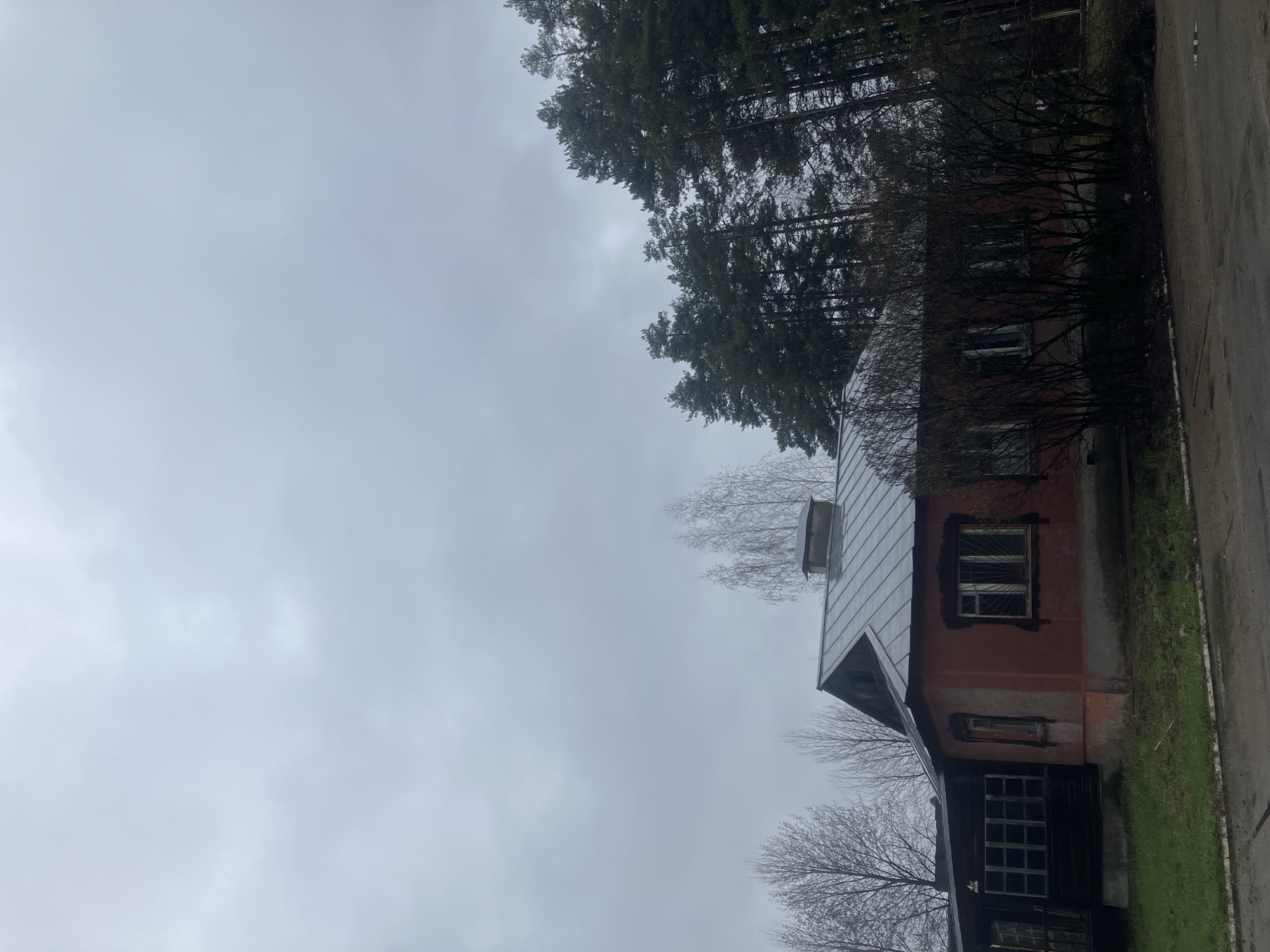 The image shows a house in Noviy, Russia. It is a two-story building with a metal roof. The house is surrounded by trees and a cloudy sky. The house appears to be old.