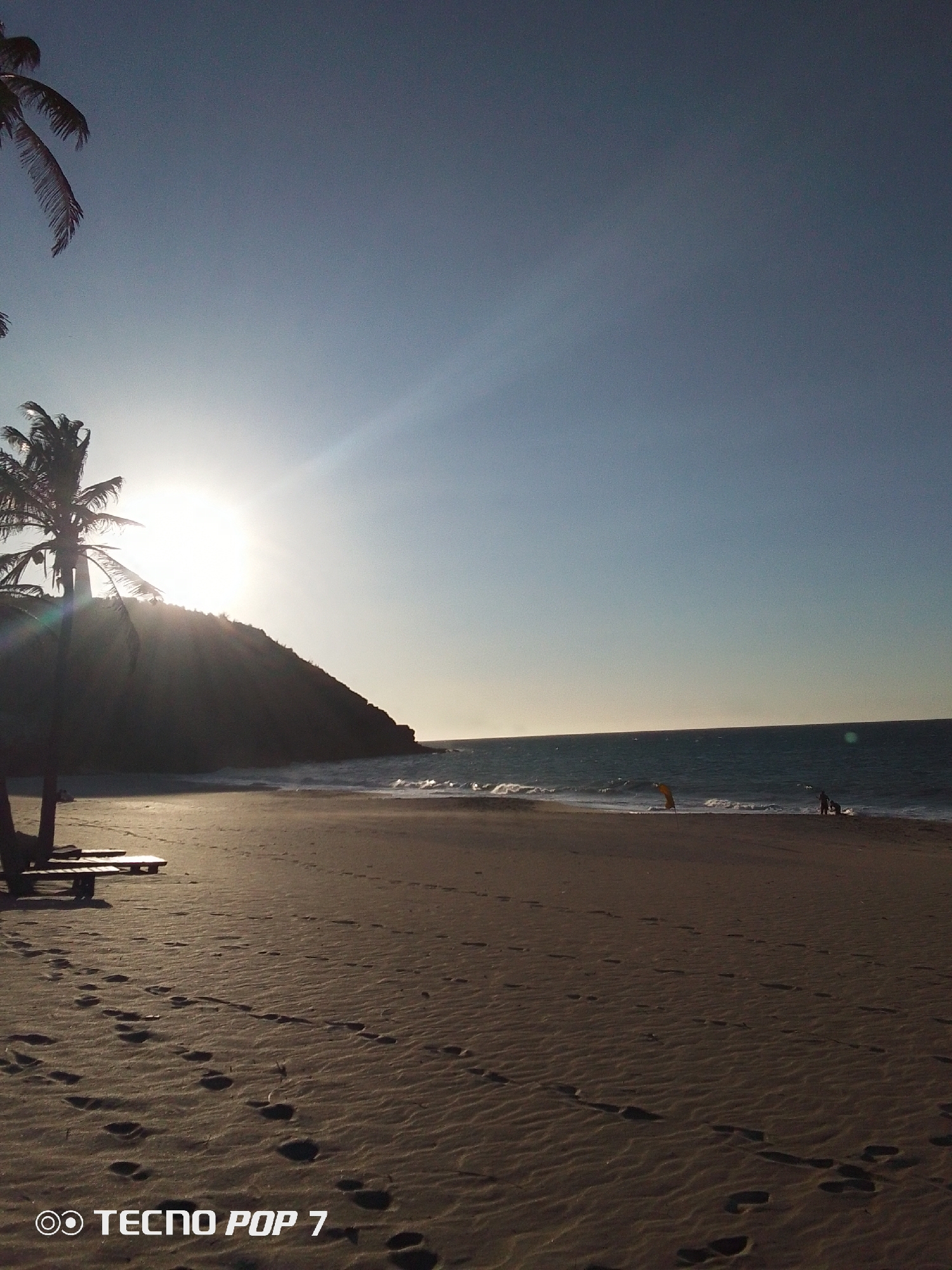 The image depicts a beach scene in Pedro González, Venezuela. The sun is setting over a prominent, verdant hill that juts into the ocean. In the foreground, there is a sandy beach with visible footprints leading toward the water. There is a wooden structure that seems to be beach furniture to the left. In the water, there is a kitesurfer and two other individuals. The sky is clear, transitioning from a light blue to a darker blue towards the top.