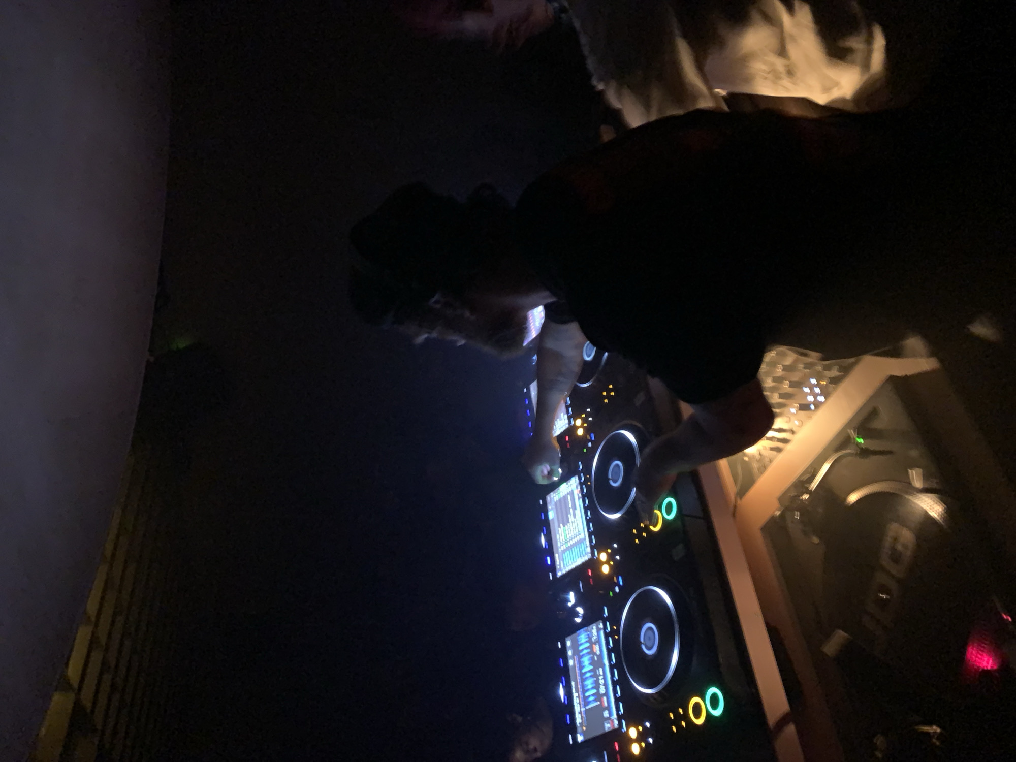 A DJ is performing on a pair of CDJ decks at an event. The location is in Tel-Aviv, Israel. The DJ wears a dark shirt and a hat. CDJs are illuminated with colorful lights. The venue is dimly lit.
