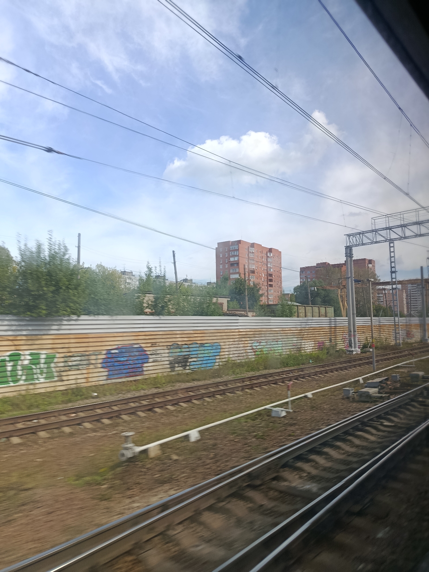 The image shows a view from a train window, looking out at a railway line and the surrounding urban landscape. In the foreground, two sets of train tracks run diagonally across the frame. A rusted metal fence, covered in graffiti, separates the tracks from a line of trees and several apartment buildings in the background. The sky is partly cloudy, with visible power lines crisscrossing the upper portion of the image. The scene appears to be located in Pavlovsky Posad, Russia.