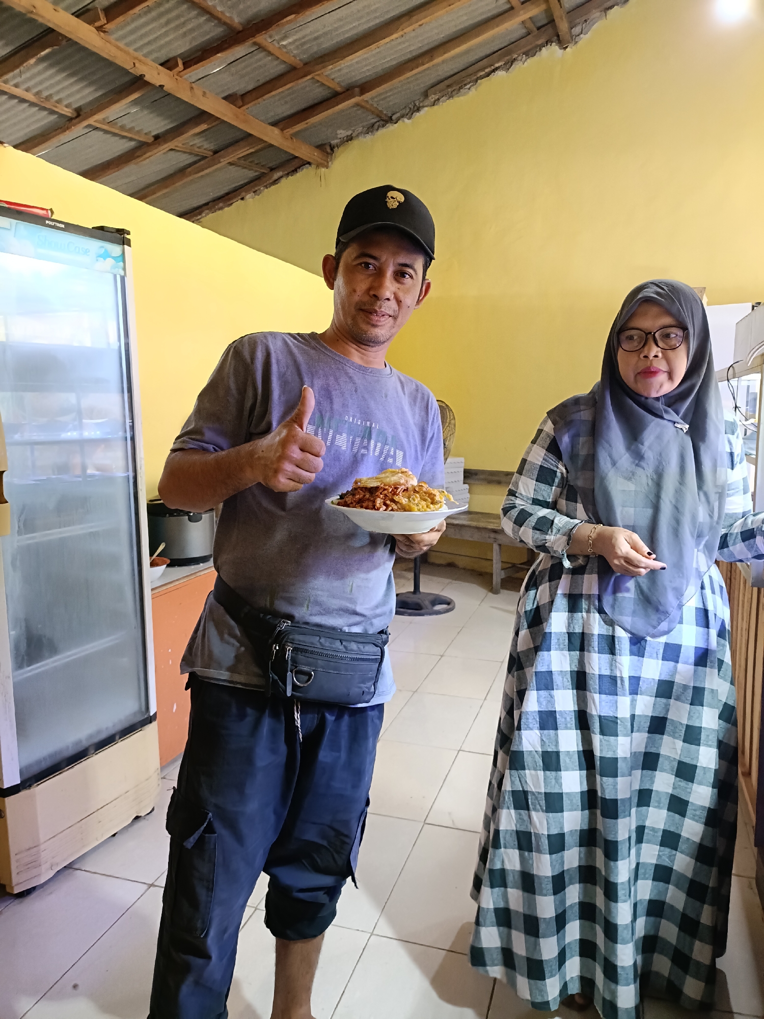 A casually dressed man in a grey t-shirt, black baseball cap, and dark cargo shorts stands in front of a yellow wall and a commercial refrigerator. He is holding a plate of food and giving a thumbs-up gesture. To his right, a woman wearing a grey hijab and a checkered dress stands near a wooden counter. The room appears to be a food establishment, possibly in Pordapor, Indonesia, with exposed ceiling beams and simple furnishings.