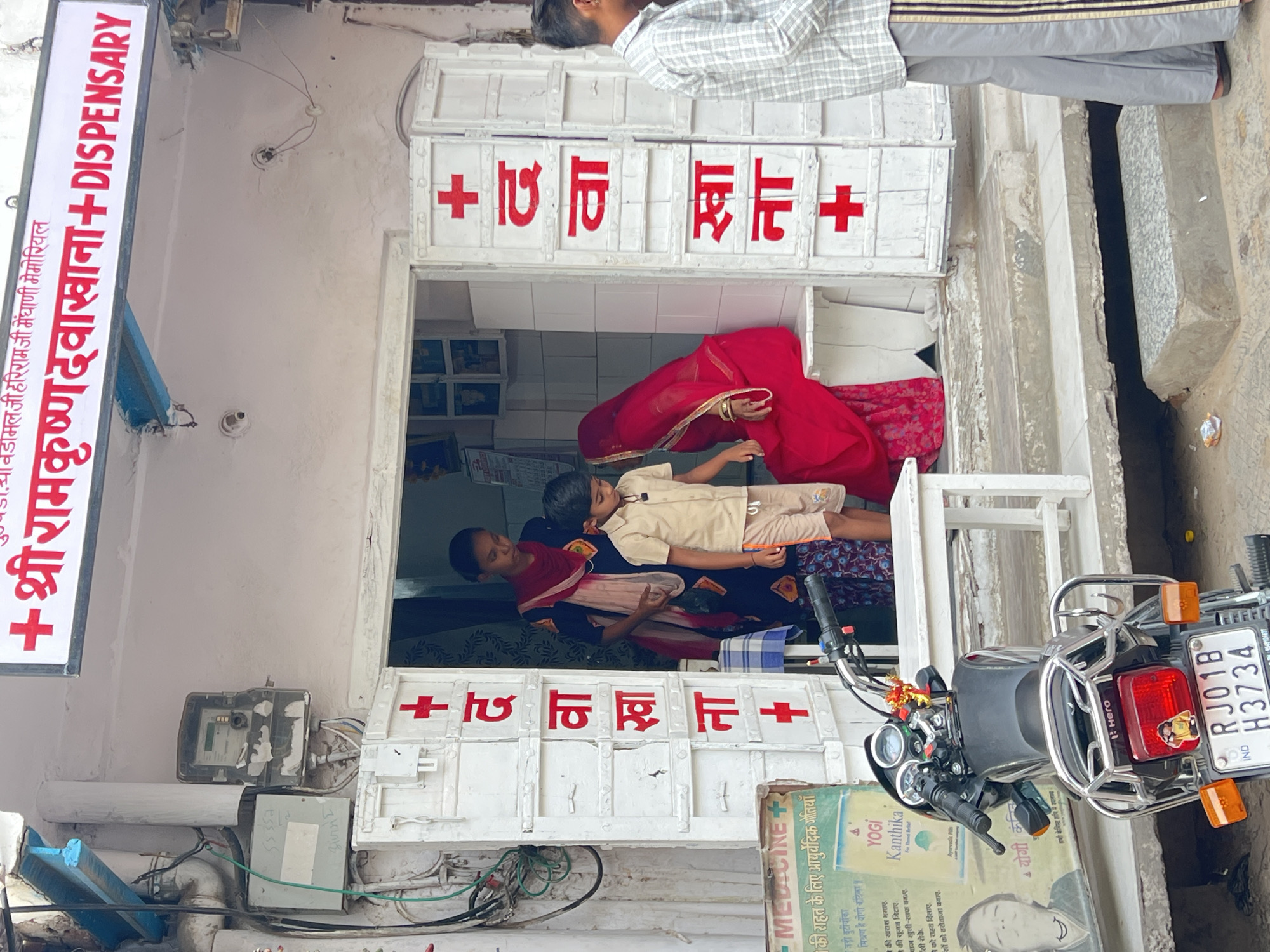 This image shows the entrance to a pharmacy in Pushkar, India. Above the entrance, a red and white sign reads "+ श्री रामकृष्ण दवाखाना + DISPENSARY." The entrance itself is framed by white doors with red plus signs and Hindi text. Inside, a young woman in a dark blue and red outfit with a red headscarf stands while a young boy in a tan shirt and shorts looks down. A woman in a bright red sari is seated to the right of the entrance. A motorcycle is parked in front of the pharmacy, and to the left, a poster advertising medicine is visible. The building appears to be made of concrete with a light-colored stucco exterior.