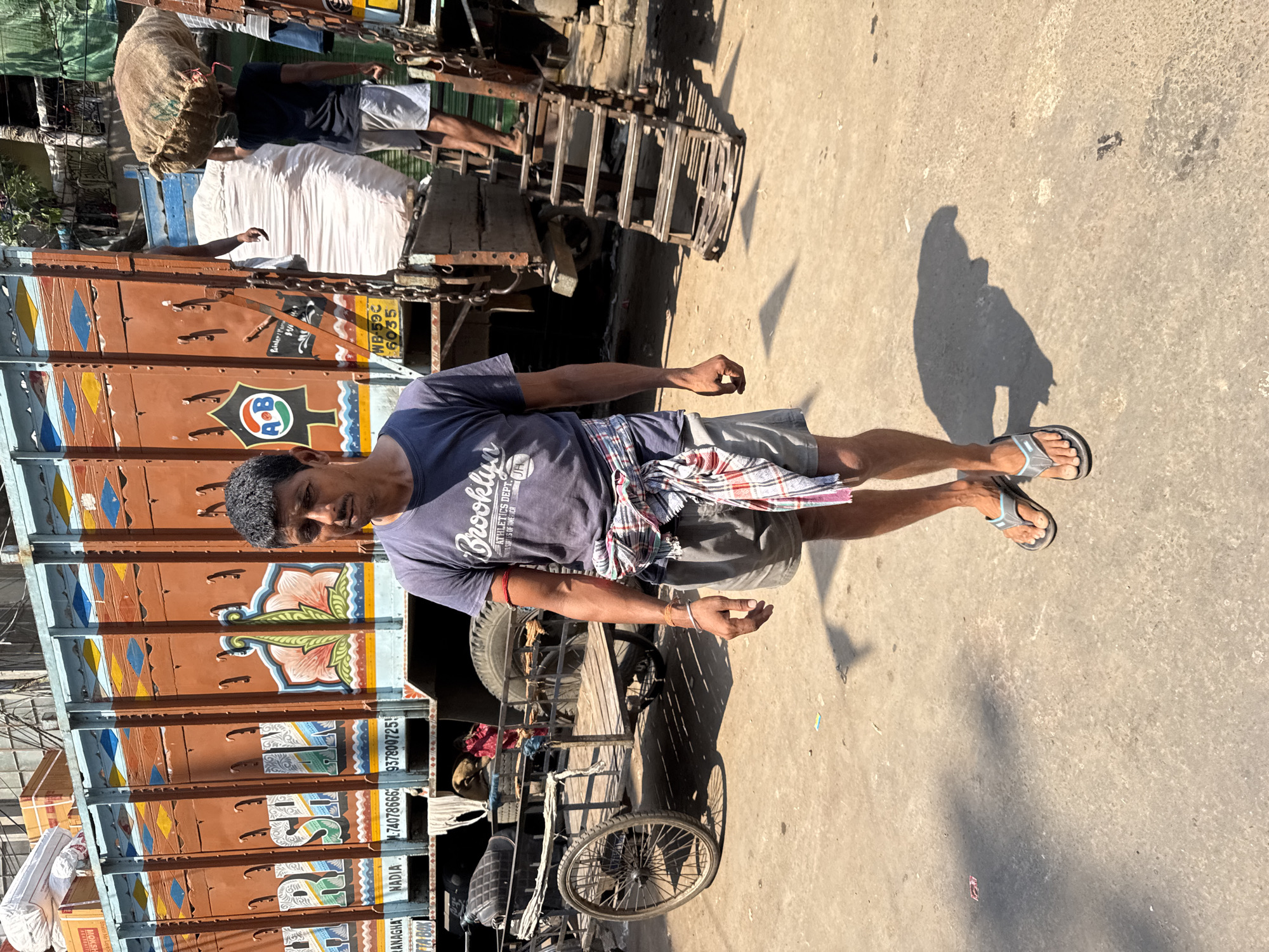 The image shows a man standing in a Kolkata street. He is wearing a blue t-shirt with "Brooklyn Athletics Dept. United States of America" printed on it, grey shorts, and blue and grey flip-flops. A patterned cloth is tied around his waist. He stands beside a brightly painted truck. A bicycle is parked in front of the truck. The ground is concrete, and the scene is bathed in sunlight. The license plate on the truck says "WB-59C 6035". There is text on the side of the truck that includes the words "RANAGHA", "NADIA", and a phone number.