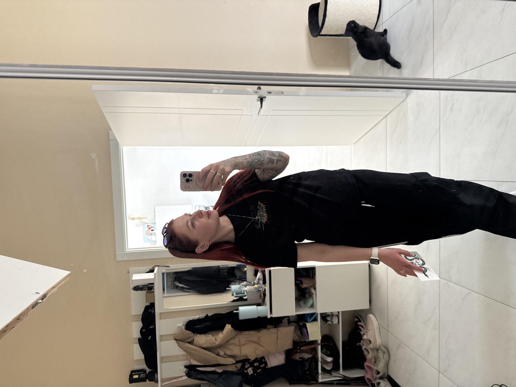 A red-haired woman is taking a selfie in front of a mirror. She is wearing a black t-shirt and dark pants. Her right arm has a visible tattoo. She is holding a phone in her left hand and a piece of paper in her right. In the background, there is a white shelf unit with various items stored on it, including clothing, shoes, and other personal belongings. There is a window above the shelf unit and a white door in the background of the mirror. A black cat is near a cat tree on the upper right. The room appears to have white tiled floors and light-colored walls. The location is unspecified, and no information indicates it is in Rostov-on-Don, Russia.