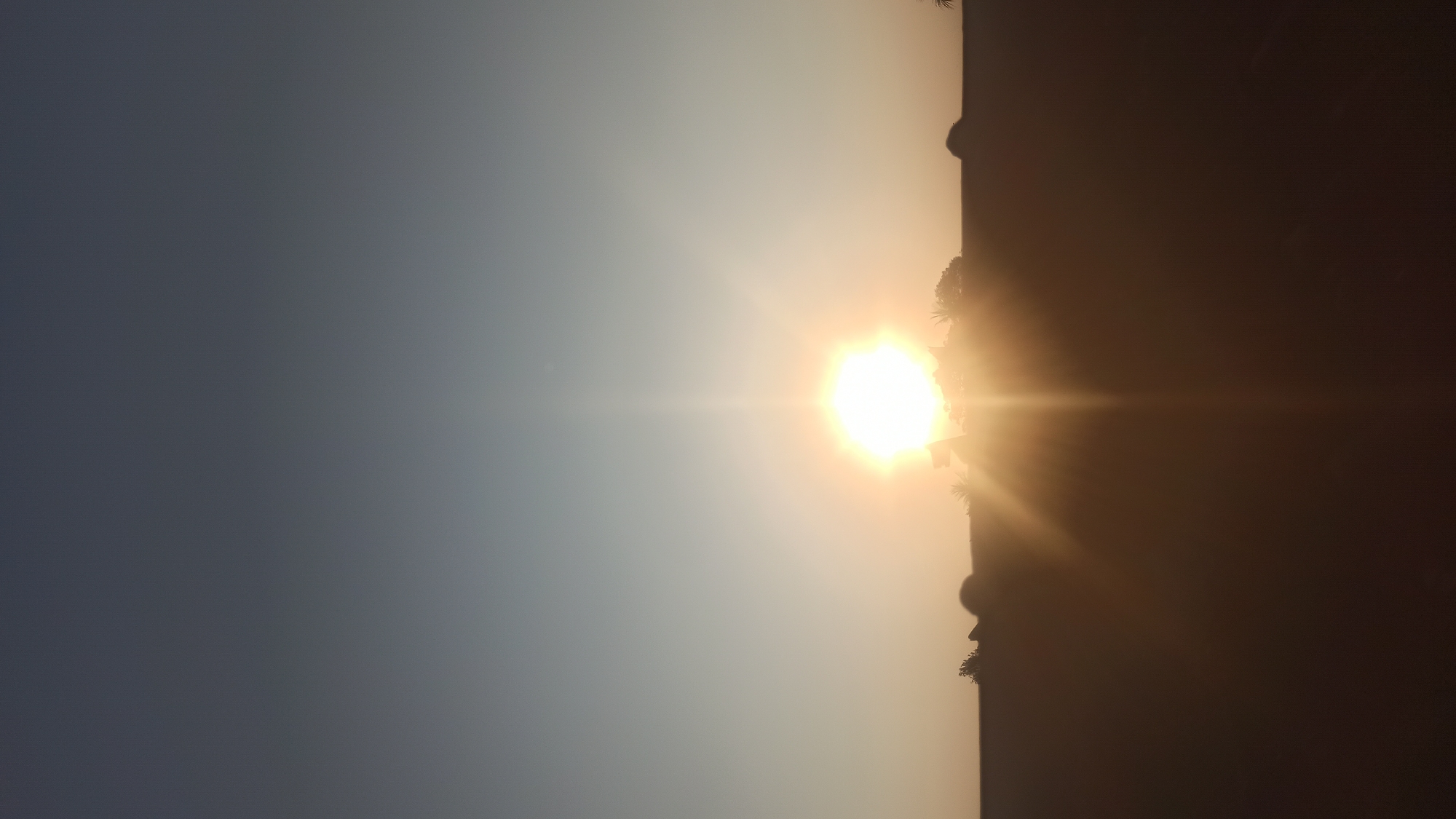 The image captures a sunset in Tuba, Indonesia, where the sun is low on the horizon, casting a warm glow. Silhouettes of trees and possibly some small structures are visible against the bright light. The sky transitions from a deep blue at the top to lighter shades of orange and yellow near the horizon. The overall scene suggests a peaceful, natural setting.