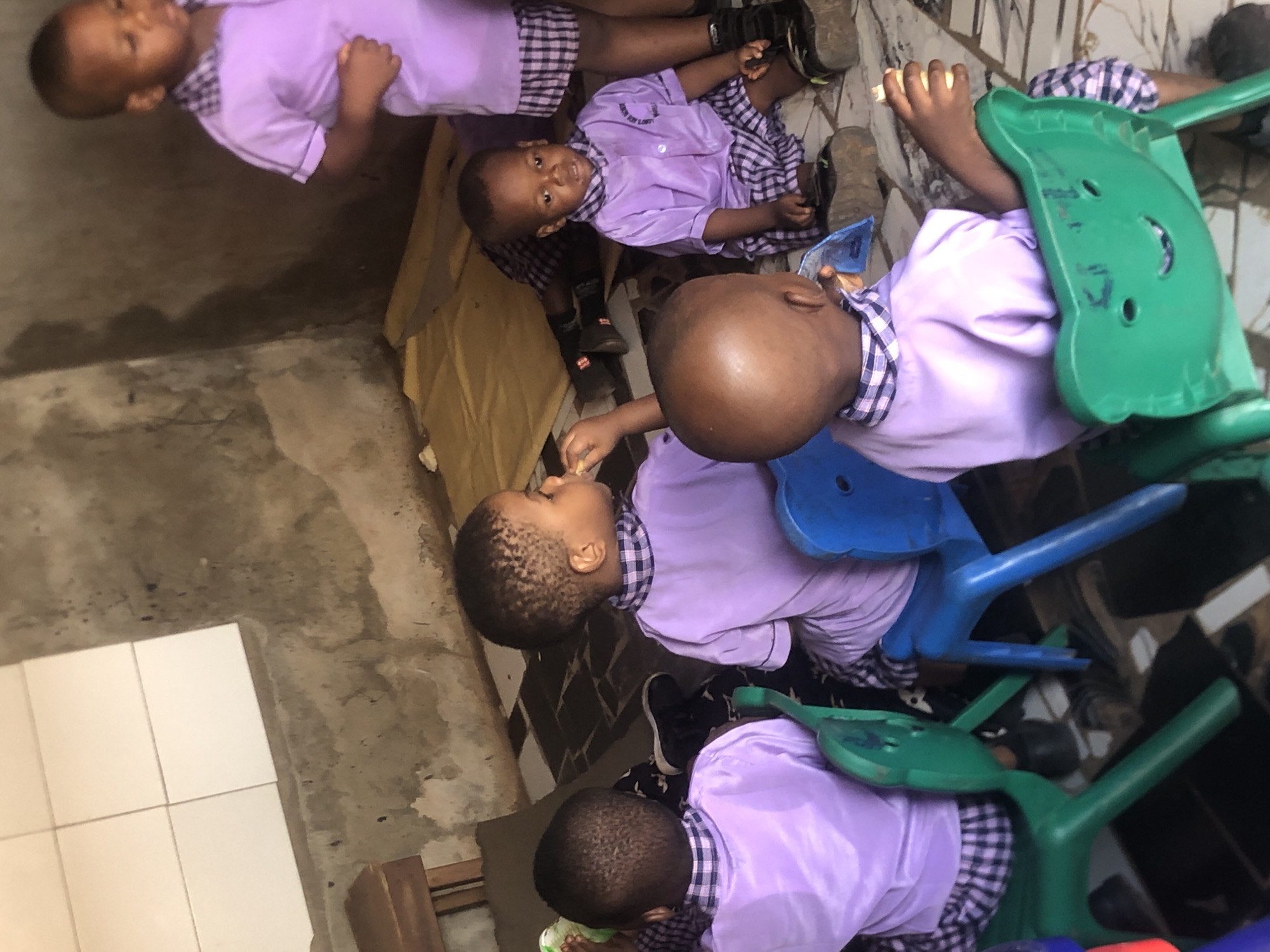 This image, taken in Ukwu-Abwa, Nigeria, shows several young children, likely toddlers or preschoolers, in what appears to be a classroom or daycare setting. The children are uniformly dressed in light purple shirts with checkered (possibly purple and white) collars and cuffs, paired with matching checkered skirts or shorts.
The children are seated on small, colorful plastic chairs, with blue and green chairs visible in the frame. The primary activity appears to be a meal or snack time; a child in the foreground is seen bringing something to their mouth to eat. Another child in the mid-ground looks directly at the camera with a gentle smile, while others are focused on their food or looking around.
The setting features concrete walls and floor, with a section of white tiling visible. On the collar of one child's uniform, a small patch is visible, bearing text that appears to read "JOY RAY SCHOOL" or a similar school name, indicating their educational institution. The overall scene depicts a calm and typical daytime moment in a children's learning environment.