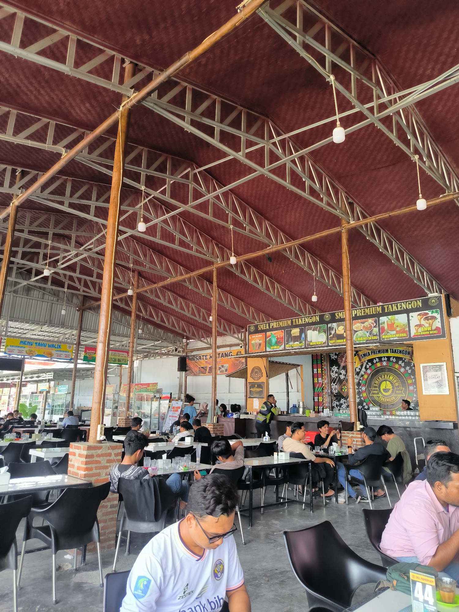 The image depicts the interior of a bustling restaurant or food court in Nunangantara, Indonesia. The space features a high, open ceiling with exposed metal framing and a dark red, textured roof covering. Wooden pillars support the structure, and several light fixtures hang from the ceiling.
Numerous tables and black chairs are arranged throughout the area, occupied by a diverse group of people. Most individuals appear to be dining or socializing. In the foreground, a young man wearing glasses and a white, purple, and black patterned shirt is seated at a table, looking down. Other patrons are visible in the middle ground and background, some interacting with each other, while others are engrossed in their phones or meals.
Various signs and banners are displayed, including "MENU TAKU WALIK" and "SMEA PREMIUM TAKENGON," suggesting food and beverage offerings. A counter area with food displays and staff can be seen in the background. The overall atmosphere appears to be casual and active, indicative of a popular eatery. The flooring is a dark, possibly concrete or tile, material.