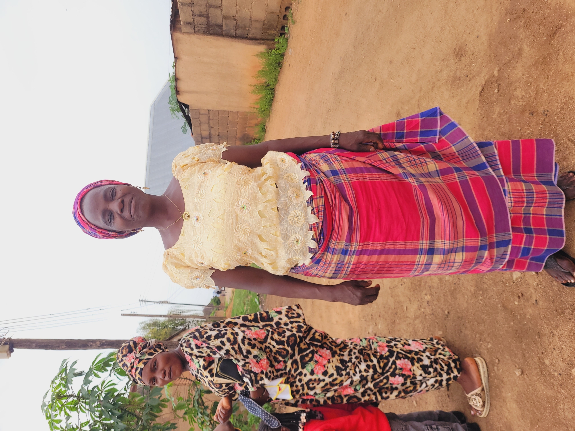 The image depicts two African women outdoors, likely in Bukuru, Nigeria. The woman in the upper portion of the frame is wearing a light-yellow, lace-like top, a colorful plaid skirt, and a patterned headwrap. She has a bracelet on her left wrist, a necklace, and is smiling. Below her, the second woman is wearing a leopard-print dress with floral patterns, a matching headwrap, and sandals. Part of a red bag and grey clothing are visible at the bottom of the frame, indicating a third person may be partially visible. The background shows a dirt ground, a building with brick or block construction, and some vegetation. A pole with electrical wires is visible in the lower part of the frame. The sky is overcast.