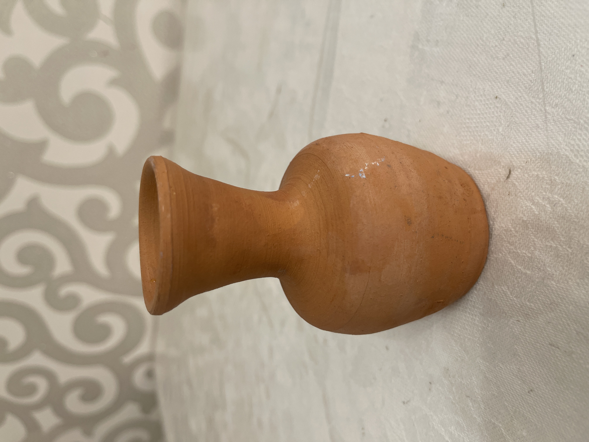 An earthenware vase with a flared rim sits on a textured white tablecloth. The vase is reddish-brown and has a rounded body that tapers to a narrow neck. The background is a wall with a light-colored, swirling pattern. The lighting is soft and natural, casting subtle shadows. The image is focused on the vase, with the background slightly blurred. There are no people or any other objects visible in the scene. The location is not specified as being in Bishkek City, Kyrgyzstan.