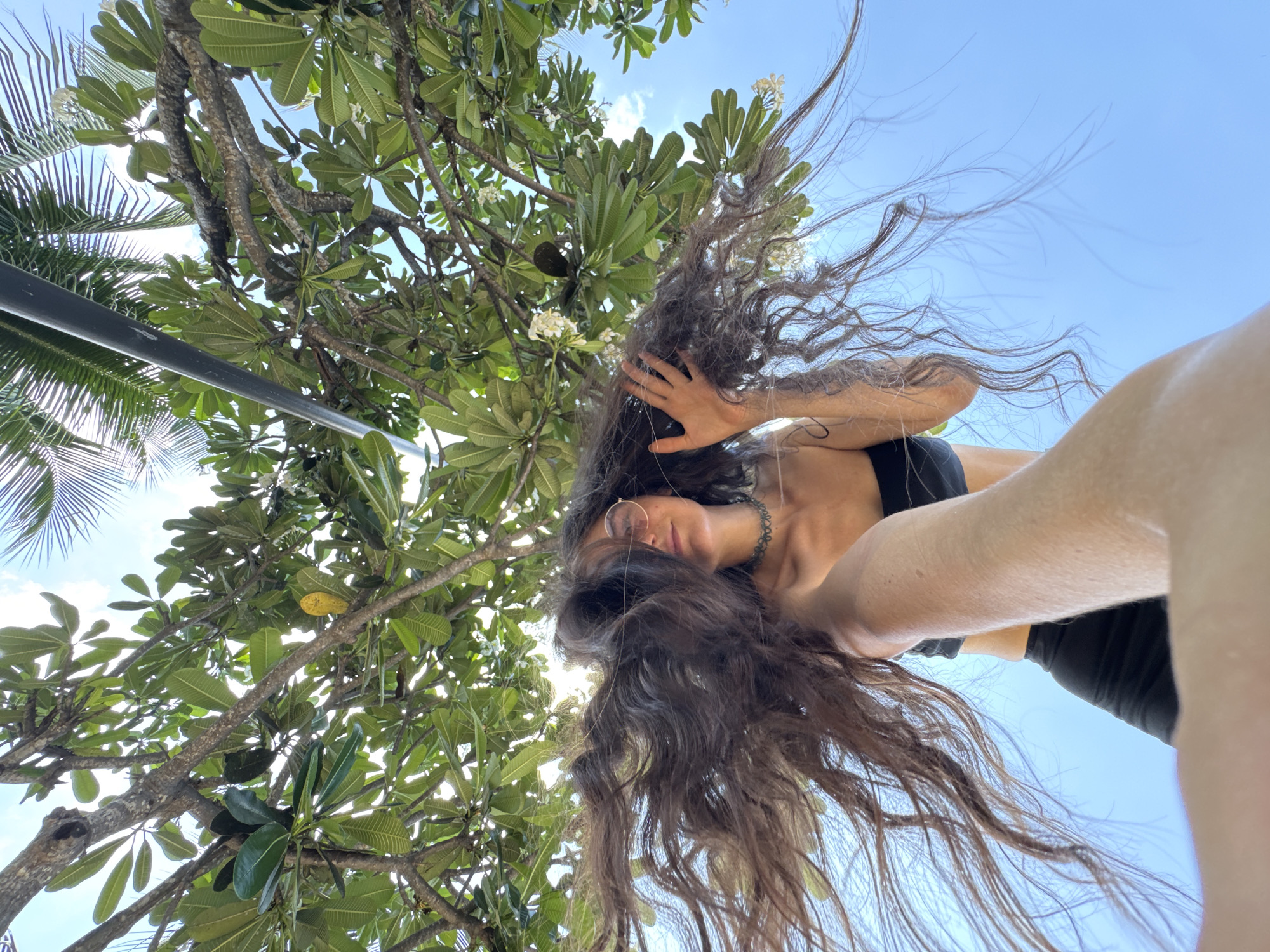 A low-angle selfie, taken in Phu Thuy Ward, Vietnam, depicts a person with long, dark brown hair splayed dramatically outwards and upwards, obscuring much of their face. The individual wears a black strapless top and dark bottoms. Round, light-tinted sunglasses cover their eyes, and a dark patterned choker necklace is visible around their neck. The person's right hand (viewer's left) is raised near their head, fingers splayed. Their left forearm and hand extend into the bottom foreground, out of focus, indicating they are holding the camera. Above the person, a dense canopy of green tropical tree leaves fills the upper frame, featuring large-leaved trees with white flowers and one yellow leaf, alongside visible bare branches and palm fronds on the upper right. A dark grey horizontal pole is present in the upper center-right. The background consists of a bright blue sky with scattered white clouds.