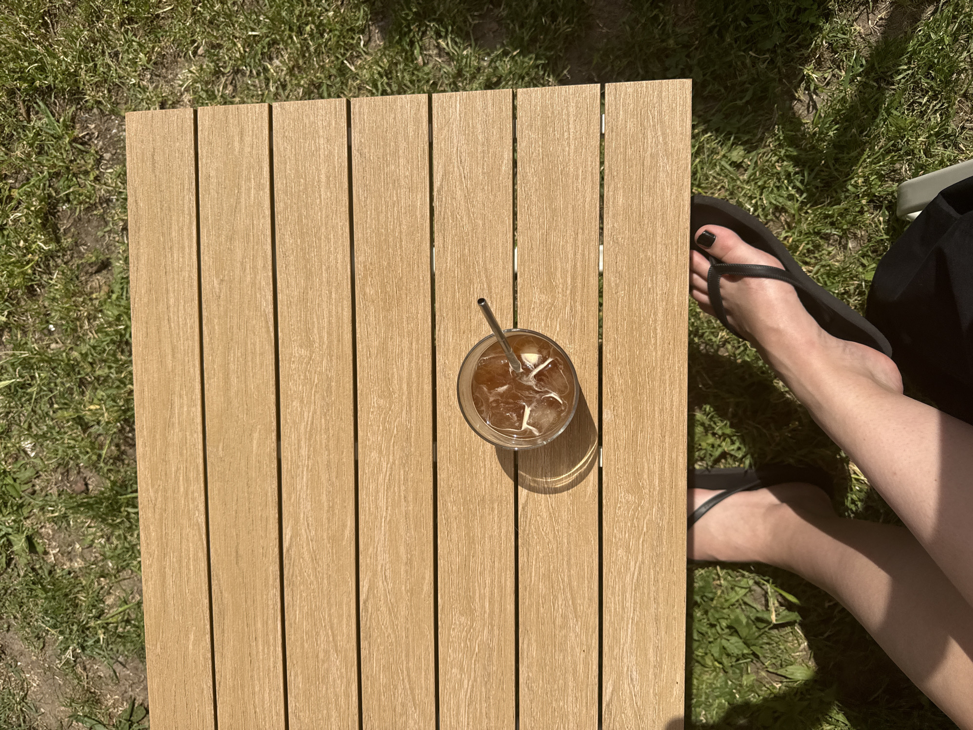The image shows a wooden table with a glass of iced coffee and a metal straw on top, with a pair of feet wearing black flip-flops visible to the side. The table is outdoors, positioned on grass. The person has black nail polish on their toes and is wearing dark clothing. The scene appears to be taken outdoors during the daytime. No clear indicators point to Belgrade, Serbia.