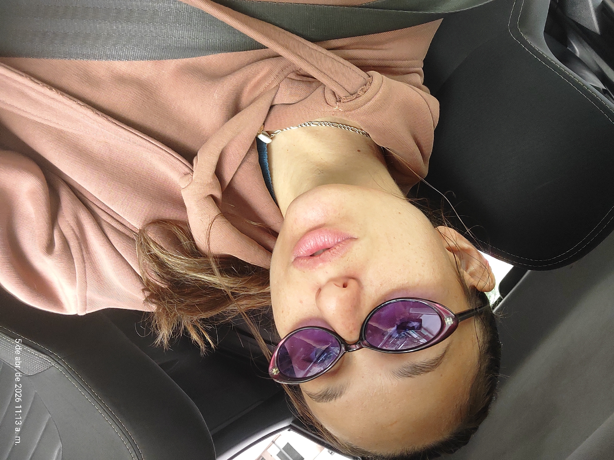 A woman is sitting in the passenger seat of a car, wearing a pink hoodie and purple sunglasses. She has a silver necklace and her hair is brown with blonde highlights. She is wearing a seatbelt. The car seat is black. The timestamp "5 de abr. de 2026 11:13 a. m." is visible in the lower left corner. The image implies the photograph was taken on April 5, 2026, at 11:13 a.m. The location is suggested to be Buenos Aires, Argentina. Buildings are visible through the sunglasses.