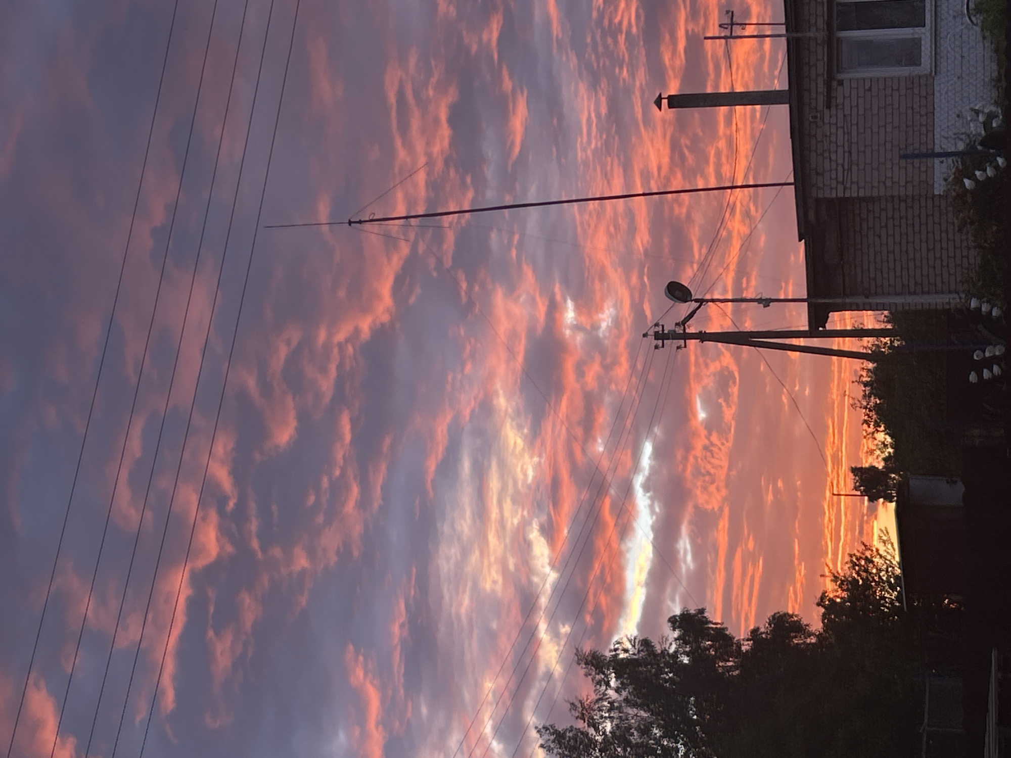 The image captures a sunset over a rural area in the Zabalotski Sel'ski Savet, Belarus. The sky is filled with dramatic clouds illuminated by the setting sun, displaying vibrant hues of pink, orange, and purple. Several power lines stretch across the sky. In the foreground, there is a building with a brick facade, a chimney, and a window. A utility pole with a light fixture and other wires is visible to the left of the building. Trees and other vegetation are present in the lower portion of the image, contributing to the rural atmosphere. The overall scene depicts a serene evening in a Belarusian countryside.