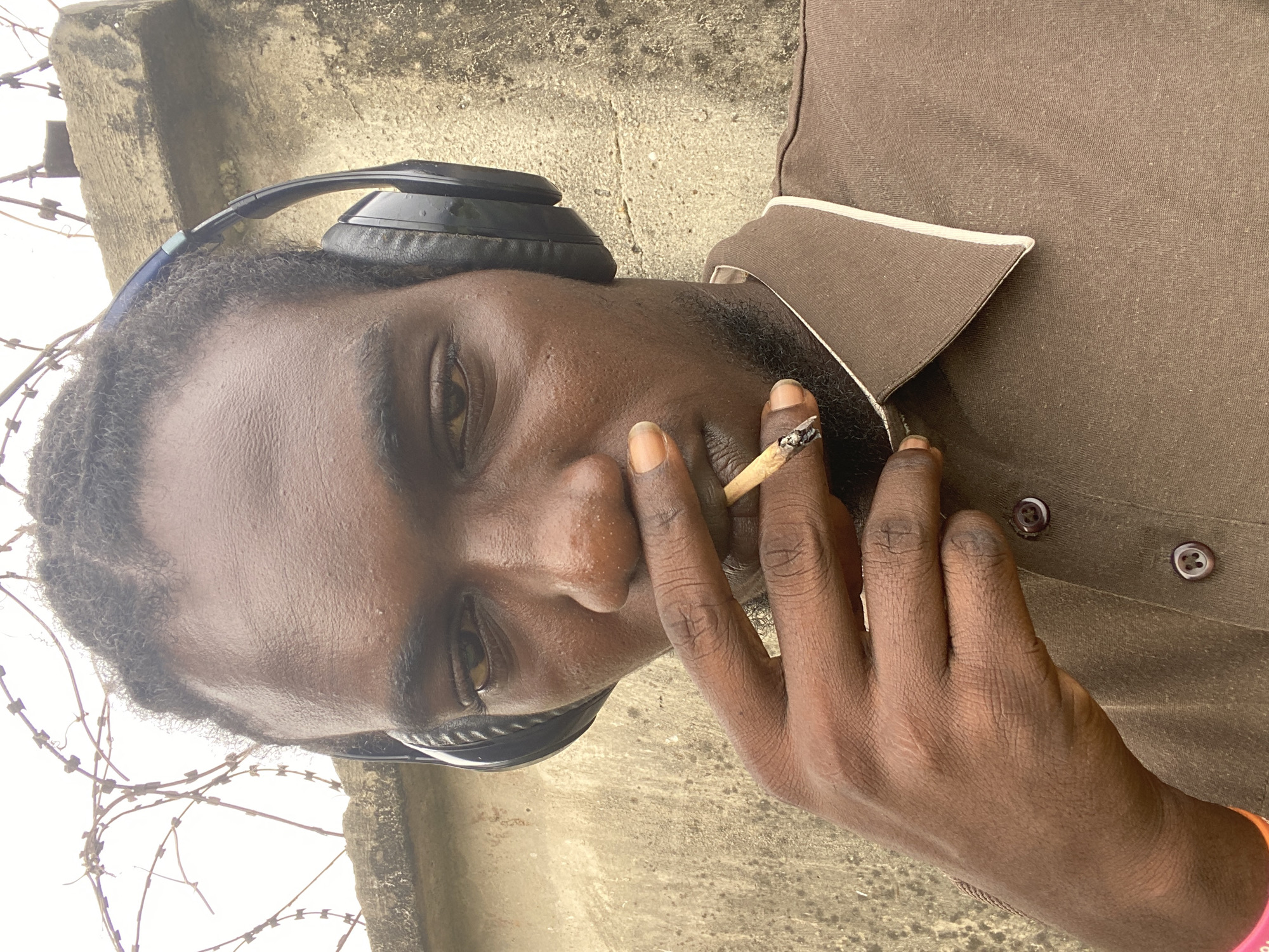 A man with dark skin and short, dark hair sits with headphones on his head, holding a lit cigarette in his right hand. He is wearing a brown shirt with a white-trimmed collar. He is positioned in front of a concrete wall topped with barbed wire. The location is suggested to be Lagos, Nigeria.