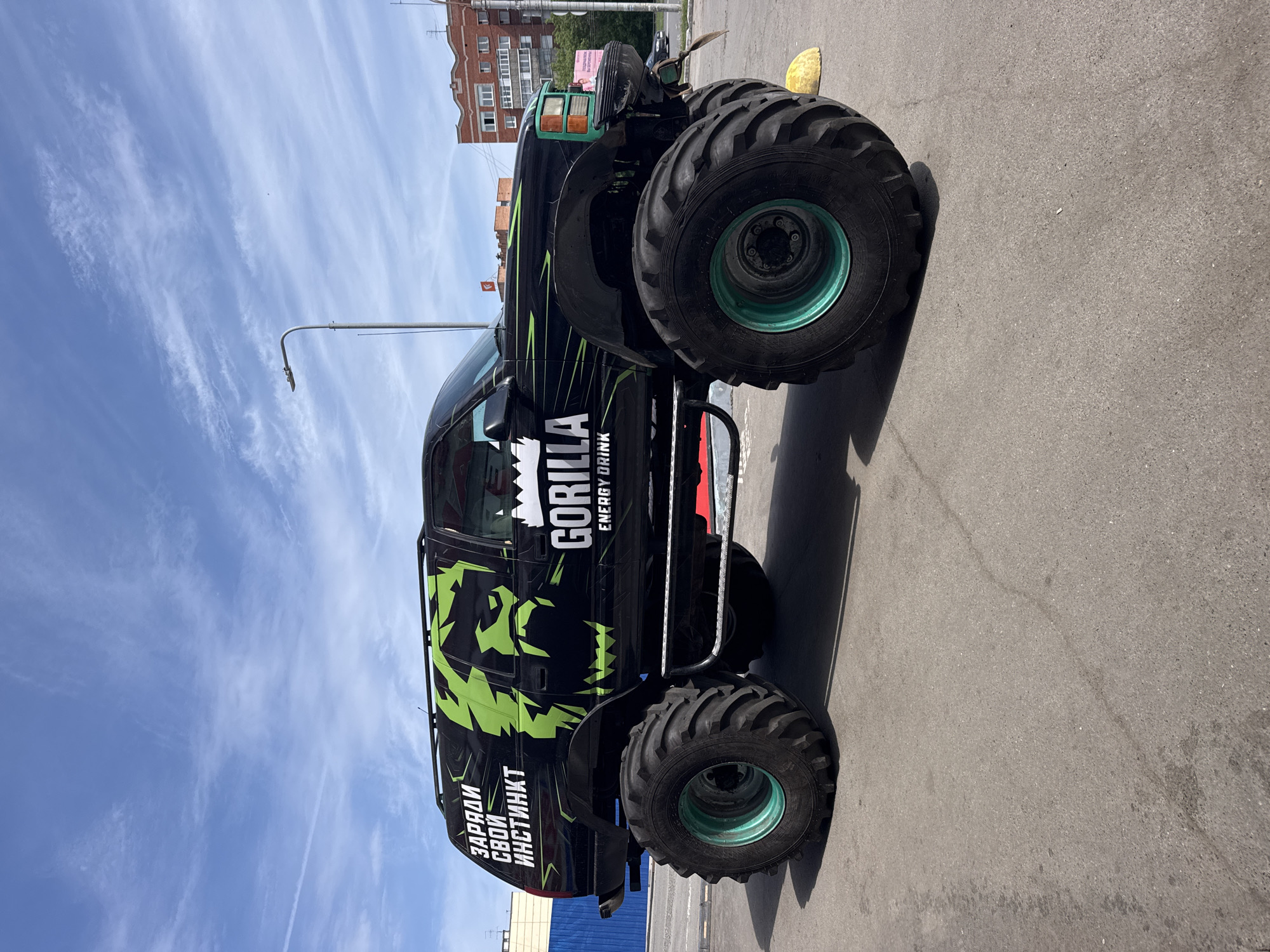 The image shows a large, black monster truck with green accents and the "Gorilla Energy Drink" logo prominently displayed on its side. The truck is parked on an asphalt surface, likely a parking lot or street. The sky is blue with wispy clouds. In the background, there are buildings, typical of an urban environment. The writing on the side of the truck is in Russian. The image was taken in the city of Izhevsk, Russia.