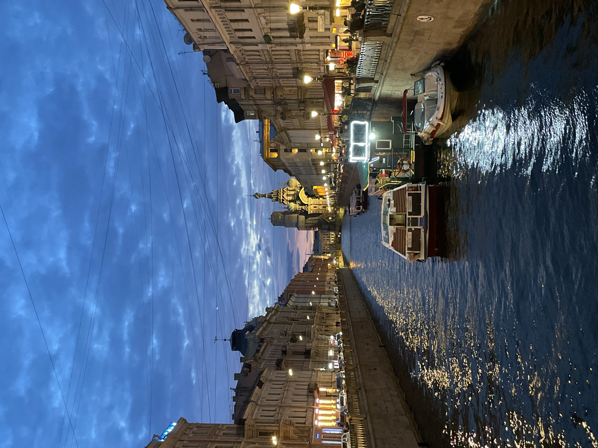 The image depicts a canal in a European city at dusk. The sky is a deep blue, with streaks of lighter blue and wisps of clouds. Overhead, a network of power lines crisscrosses the frame. On either side of the canal, ornate buildings with illuminated windows line the banks. Several boats are moored or navigating the water, some appearing to be tour boats with people on deck. Streetlights cast a warm glow on the facades of the buildings and reflect on the water's surface, creating shimmering patterns. In the distance, a prominent domed building, likely a church or cathedral, with intricate architecture stands out against the twilight sky. Cars are visible along the street on the right bank, and pedestrians can be seen walking along the sidewalks. The overall atmosphere is one of a vibrant city transitioning from day to night.