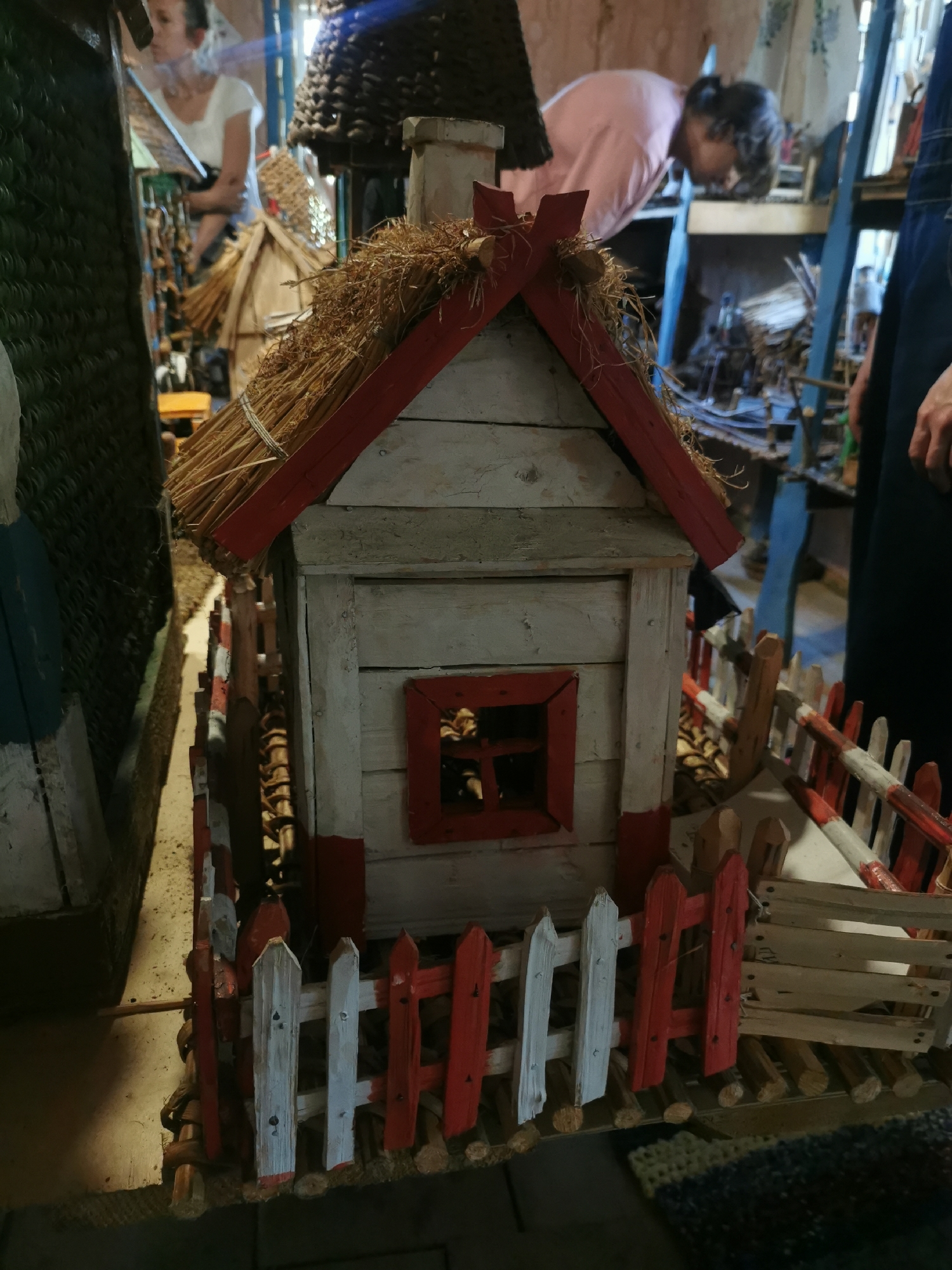 In a dimly lit indoor setting, likely a workshop or a shop displaying handicrafts, a small, weathered wooden house with a thatched roof and red trim stands prominently in the foreground. The house has a single small window framed in red. A miniature picket fence, alternating white and red, surrounds the house. In the background, partially obscured and out of focus, are two individuals. One, on the left, appears to be a woman in a white shirt, looking down at something. Another individual, on the right, is wearing a pink shirt and is also looking downwards, possibly working on something. The environment suggests a place where handmade crafts are created or sold, with various items and materials visible in the background, including baskets and what appear to be tools. The overall atmosphere is one of focused activity in a rustic, possibly traditional, setting. The location is identified as Shcharchoŭski sel’ski Savet, Belarus.