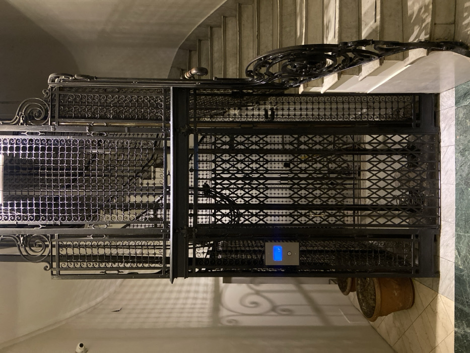 The scene depicts an antique, ornate wrought iron elevator shaft and an adjacent marble staircase within an interior building environment. The elevator, central to the image, features dark, intricate wrought iron construction with grid patterns and decorative scrollwork, particularly on its upper section. The ground-level elevator entrance is secured by a black metal collapsible scissor-gate door. A silver-toned call panel with a blue illuminated display and a round blue button is affixed to the left of the elevator door frame. Two empty terracotta pots are situated on the tiled floor to the elevator's left. To the right, a multi-tiered staircase with light-colored marble steps ascends, bordered by a dark, ornate wrought iron balustrade topped with a wooden handrail. The flooring at this level consists of white and green marbled tiles. The walls are light-colored, and a white security camera is mounted high on the upper-left wall. Shadow patterns from the elevator's ironwork are visible on the left wall. No individuals are present. This architectural style is typical of early 20th-century buildings in historical districts of Buenos Aires, Argentina, such as Recoleta.