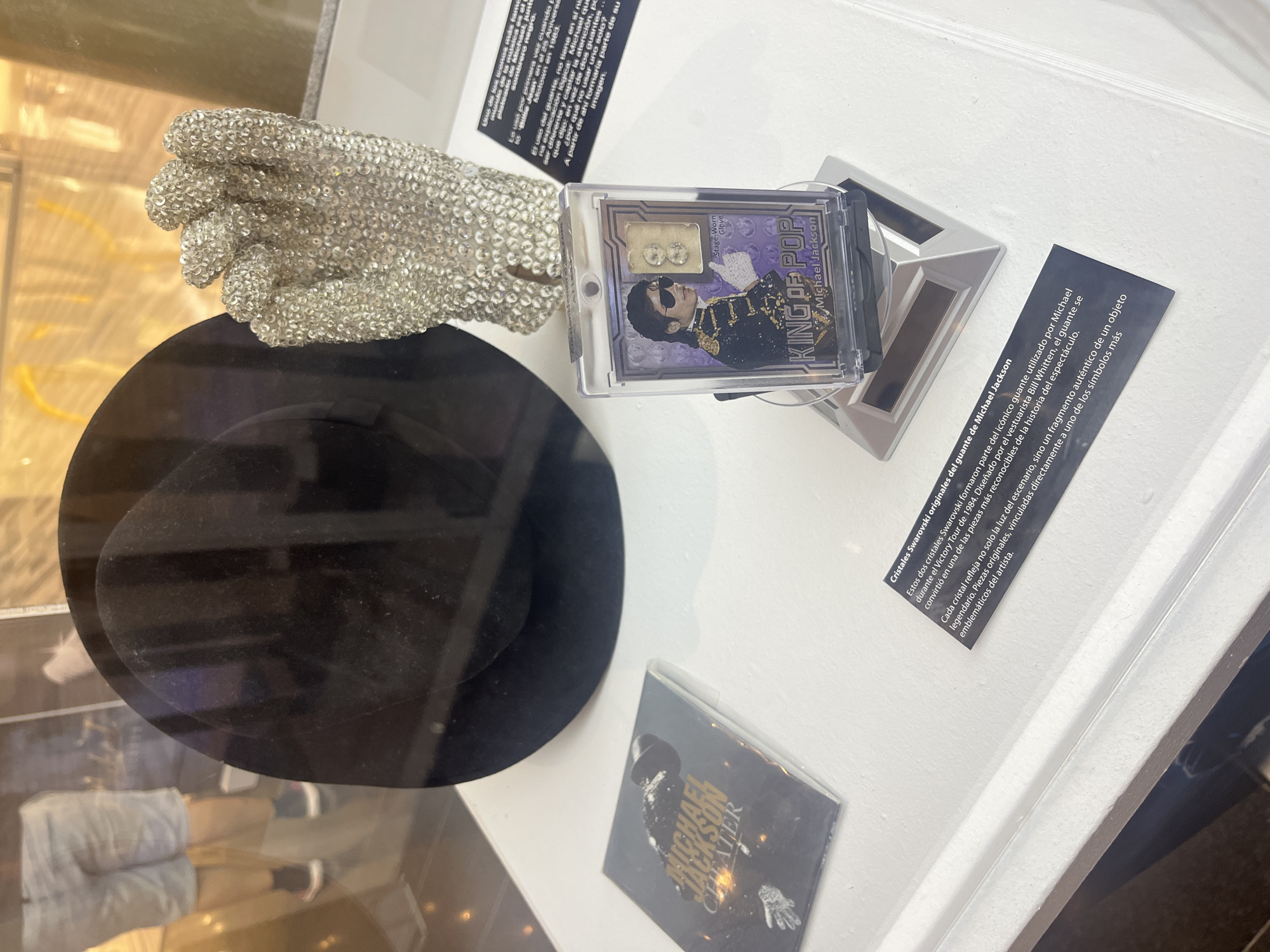 The image displays a collection of Michael Jackson memorabilia inside a brightly lit display case, likely in an exhibition or museum in Las Palmas, Spain. The central objects are a sparkling, rhinestone-covered glove, characteristic of Jackson's iconic stage attire, and a black fedora hat.
To the right of the glove, a clear acrylic case holds a collectible item featuring an image of Michael Jackson in sunglasses and a military-style jacket, labeled "KING OF POP Michael Jackson". Inside this case, two small, clear, possibly crystal fragments are visible, identified by text as originating from a "Stage-Worn Glove". Below the fedora, a small, square item with "MICHAEL JACKSON THEATER" printed on it is partially visible.
Several informational plaques in Spanish provide context for the artifacts. One plaque explains that the two small fragments are "Original Swarovski crystals from Michael Jackson's glove," which were part of the iconic glove worn during the 1984 Victory Tour and designed by Bill Whitten. It notes that these crystals are authentic fragments of a legendary object and emblematic of the artist. Another partially visible plaque discusses the origin of Jackson's decision to wear only one glove, mentioning "Billie Jean" and "Motown in 1983", stating he sought to be unique and attention-grabbing, making the single glove a signature part of his image.
Reflections on the display case glass show the lower half of a person, including legs in shorts and sneakers, indicating an indoor setting with visitors. The white background of the display case accentuates the items, creating a clean exhibition environment dedicated to the legacy of the pop superstar.