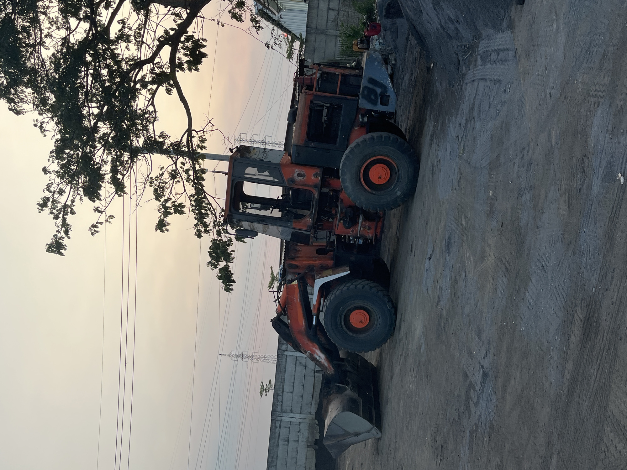This is a photograph of an orange and grey front-end loader parked on a dirt lot in Kedungcangkring, Indonesia. The loader is partially dilapidated, with some of its paint peeling and the cab appearing damaged. It is positioned in the center-left of the frame, with its bucket lowered to the ground. Behind the loader, a concrete wall runs horizontally across the background, and beyond that, electrical pylons and trees are visible against a pale, hazy sky that suggests either dawn or dusk. To the right of the loader, a pile of dark material, possibly coal or gravel, is present, and a small, red jerrycan is visible near the front of the loader. The ground is covered in dirt and tire tracks, indicating recent activity. The scene appears to be an industrial or construction site.