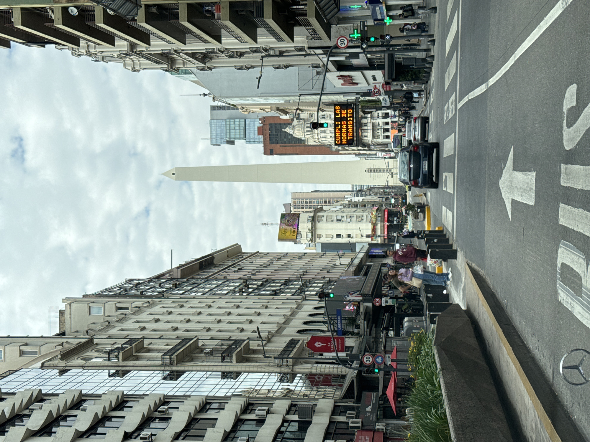 This image captures a bustling urban street scene in Buenos Aires, Argentina, looking down a wide avenue towards the iconic Obelisco de Buenos Aires, which stands prominently in the background under an overcast sky with scattered clouds. The perspective suggests the photo was taken from a vehicle, with the street and road markings in the foreground.
The street itself features multiple lanes, with a prominent white arrow painted on the asphalt indicating the direction of traffic, and a partially visible "BUS" marking. A Mercedes-Benz logo is also painted on the road surface. Cars, mostly dark-colored, are visible in the lanes, some stationary. On the right side, a yellow curb delineates a bus lane.
Along both sides of the avenue, a mix of architectural styles is present, from modern high-rise glass buildings to older structures with ornate details and balconies. Sidewalks are visible on both sides, populated by pedestrians. On the right, a small group of people, including two individuals (one in a maroon top, another in a light green top), are standing on the sidewalk near some street furniture and greenery.
Various signs and details contribute to the urban atmosphere. A bright orange LED sign displays the message "CUMPLI LAS NORMAS DE TRANSITO" (Obey traffic rules). Traffic lights showing green are visible, as are street signs indicating a "30" km/h speed limit and no parking restrictions. A red "FARMACIA" (Pharmacy) sign is also discernible above a storefront. In the distance, a large billboard with "ORO" visible suggests commercial activity. The scene depicts everyday life on a major thoroughfare during what appears to be daytime.