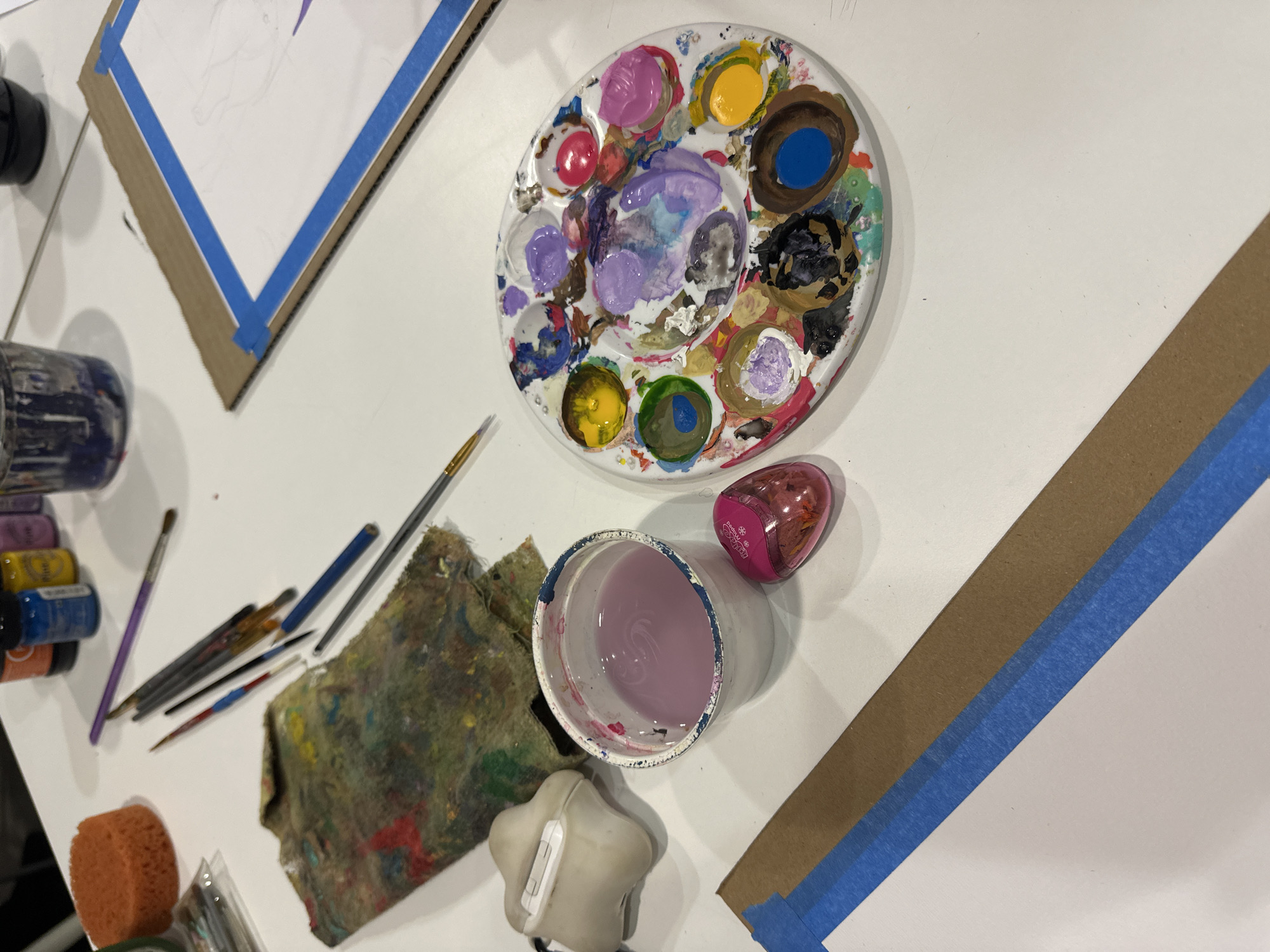 The image depicts an artist's workspace, likely indoors. Dominating the scene are a white plastic paint palette with various colors of wet paint, a white container with a light purple liquid (likely paint or cleaning water), and a pink pencil sharpener. Various paintbrushes and a sponge lie beside a paint-stained cloth. Two canvases are visible, secured with blue painter's tape to cardboard backing. Partially visible is an unlabeled row of small jars or bottles that appear to contain various paints.
There is no information available to determine the specific location in Mexico City, Mexico.