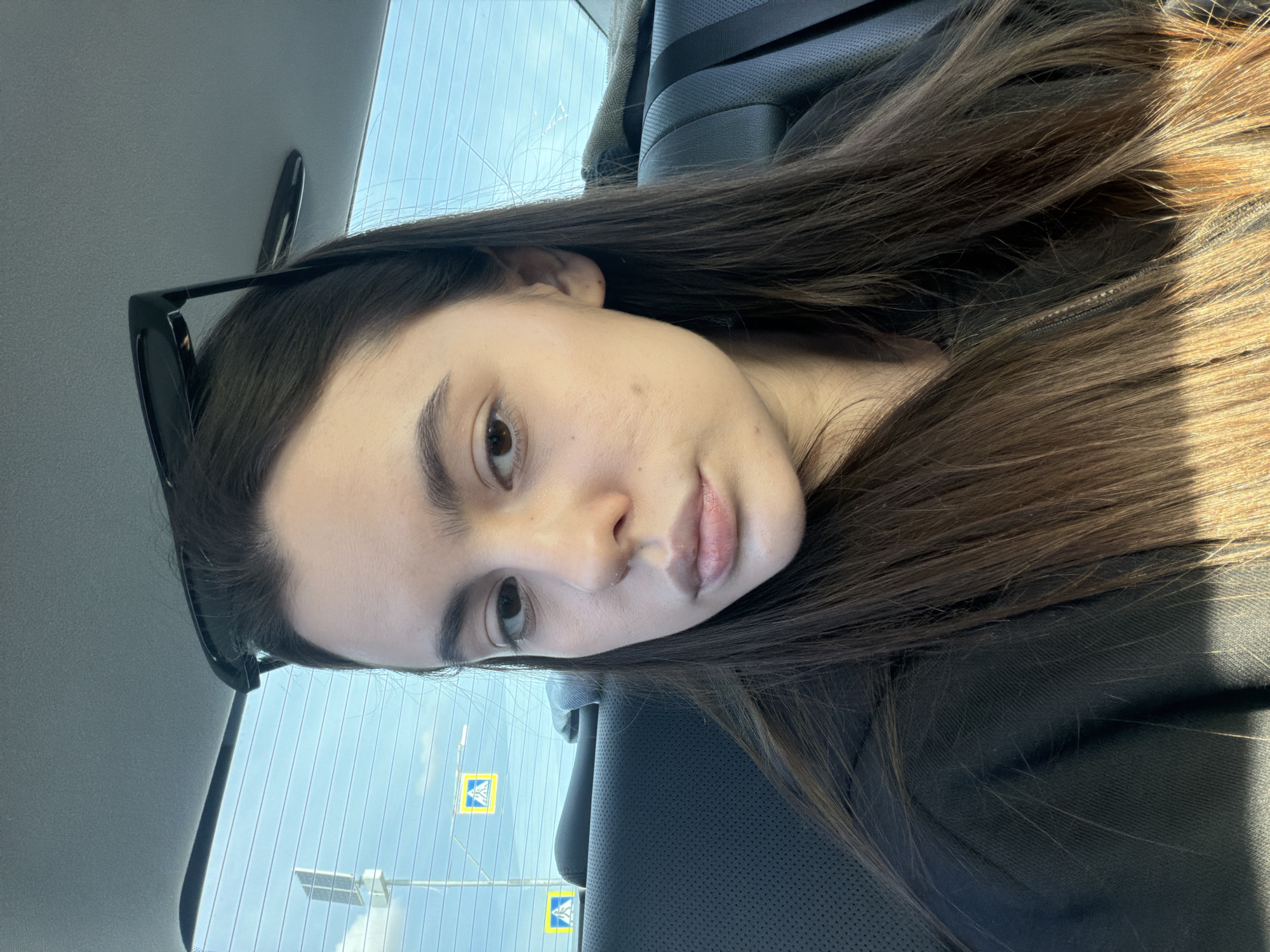 A young woman is captured in a portrait-oriented, eye-level shot while seated inside a vehicle during the daytime. She appears to be in her late teens or early twenties, looking directly at the camera with a neutral expression and slightly parted lips. Her long, dark brown hair, featuring lighter sun-kissed streaks, flows over her left shoulder and chest. She has sunglasses resting on top of her head and is wearing a dark, possibly black, top or jacket with a visible zipper detail on her right side.
The setting is the interior of a car, evidenced by the dark, textured upholstery of the seats and a seatbelt strap visible over her right shoulder. Through the car's rear window, a bright blue sky with some wispy clouds is visible, streaked with horizontal lines, likely from the defroster or reflections. Outside the window, two yellow road signs, each displaying a blue square with a white pedestrian crossing symbol, are clearly identifiable. One sign is prominent in the lower left portion of the window, with another partially visible further away. A dark pole or structure is also seen outside. The strong natural light indicates it is daytime. The location is Сосново, Russia. No discernible text is present in the image.