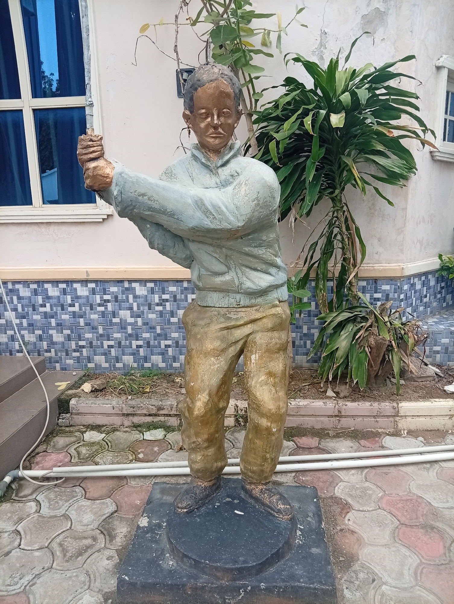A full-body statue of a young man stands outdoors in Jalingo, Nigeria. The statue is made of a material that resembles bronze for the skin and gold for the clothing and hair. The man is depicted in mid-action, possibly swinging a club or racket, with his body bent and his arms raised. He wears a light blue jacket over tan pants and black shoes. The statue is mounted on a dark, circular base, which rests on a patterned pavement made of interlocking concrete tiles in shades of pink and grey.
Behind the statue, a pale pink building with white trim is visible. To the left, a large window with blue-tinted glass reflects the surroundings. A lush green plant with long, broad leaves grows to the right of the statue, partially obscuring the building. Further to the right, another window is visible. The ground around the statue is a mix of soil, some dry grass, and more of the patterned paving. White pipes run along the ground in front of the statue, suggesting some form of outdoor utility or irrigation. The overall scene suggests an outdoor decorative element in a residential or public space. The lighting suggests it is daytime, with no strong shadows indicating a specific time. The weather appears to be clear and sunny. There is no visible text in the image.