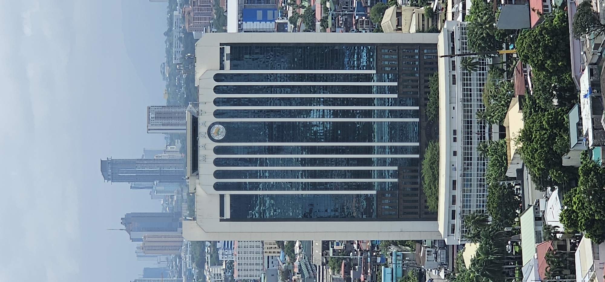 The image shows a tall, modern building with a glass facade, identified by signage as the "CITY OF MAKATI PHILIPPINES". The building is situated in a dense urban environment with other skyscrapers and lower-rise buildings visible in the background. The sky is overcast, suggesting a cloudy or hazy day. The foreground shows a lush green landscape with trees and rooftops of residential buildings. The main building features a series of vertical white columns against the blue-tinted glass windows. At the top of the building, there are arched elements and a circular emblem. The overall impression is one of a metropolitan area with a mix of modern and older architecture, characteristic of Makati City, Philippines.